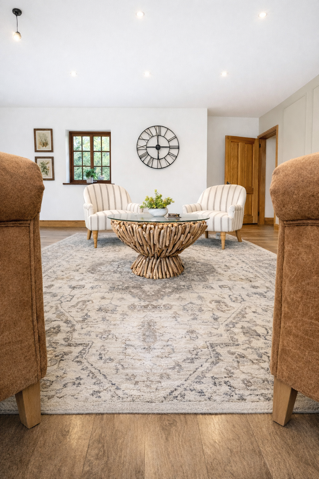 A warm contemporary living and dining space with neutral tones and natural textures. Two tan leather armchairs with cushions are positioned in a patterned rug beside a round glass and stone coffee table. Behind them, tall wooden-framed glass doors op