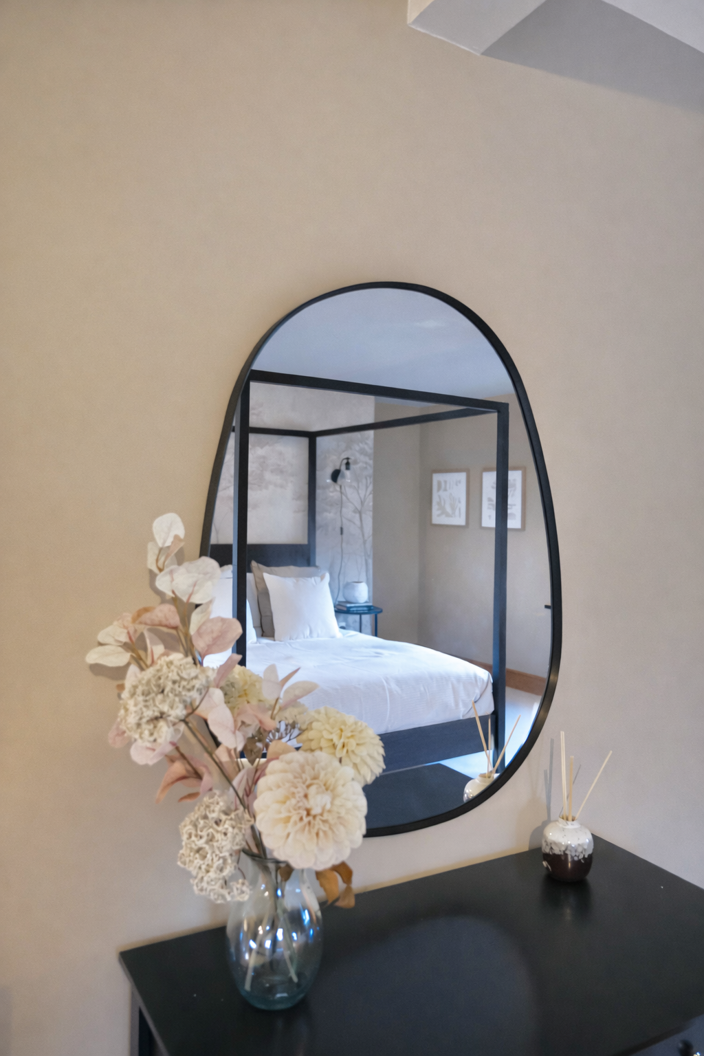 An oval shaped mirror with a thin black frame mounted on a beige wall. Mirror reflects the bedroom featuring a bed with white bedding and black four-poster bed frame. Below the mirror is a dark dresser. There is a vase with dried - neutral toned flow