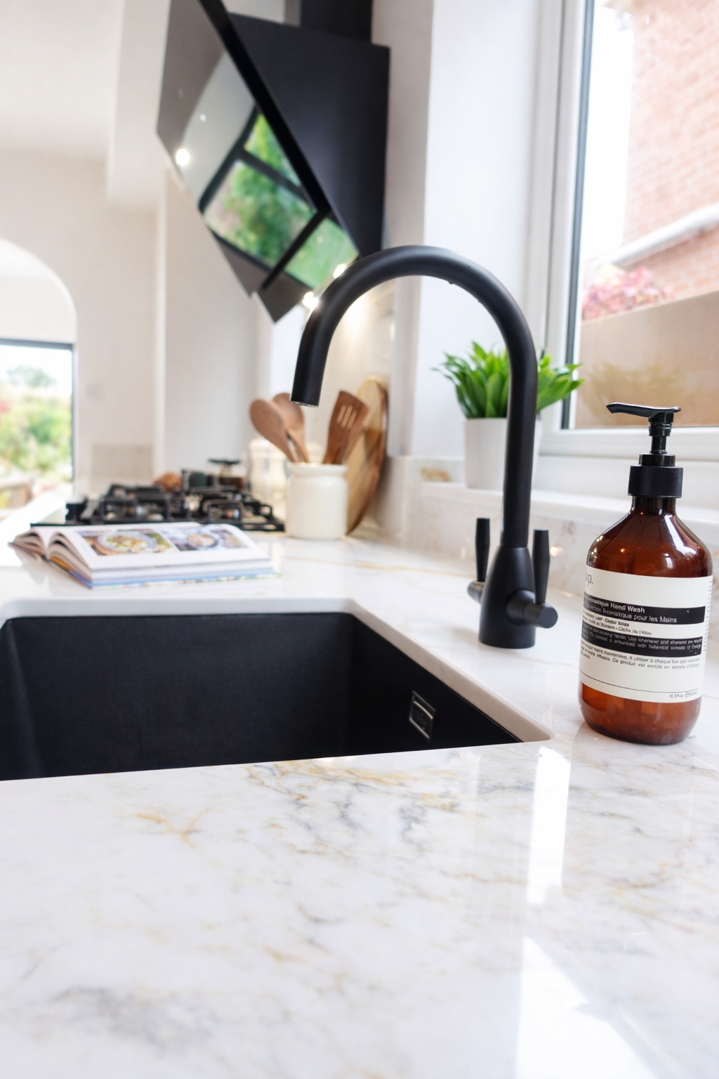 A close-up view of the kitchen’s sleek sink area, showcasing a contemporary black tap paired with a deep black inset sink set into a light marble-effect worktop. Positioned beside a bright window, the space is filled with natural light, highlighting 