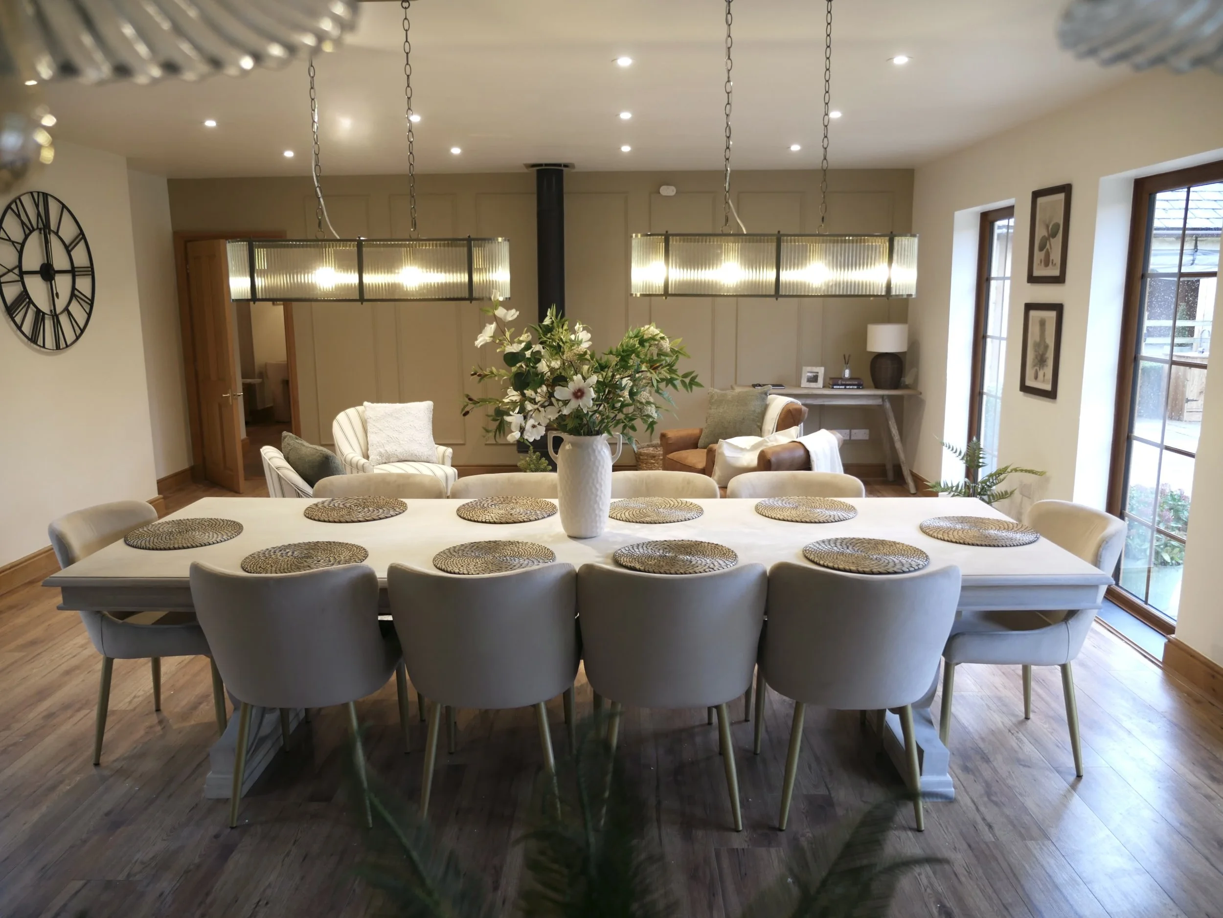A bright and airy dining space featuring a large white table surrounded by ten comfortable chairs. Woven placemats add natural texture, while a vase of fresh flowers sits at the centre, bringing life and colour to the table. A pendant light hangs ove