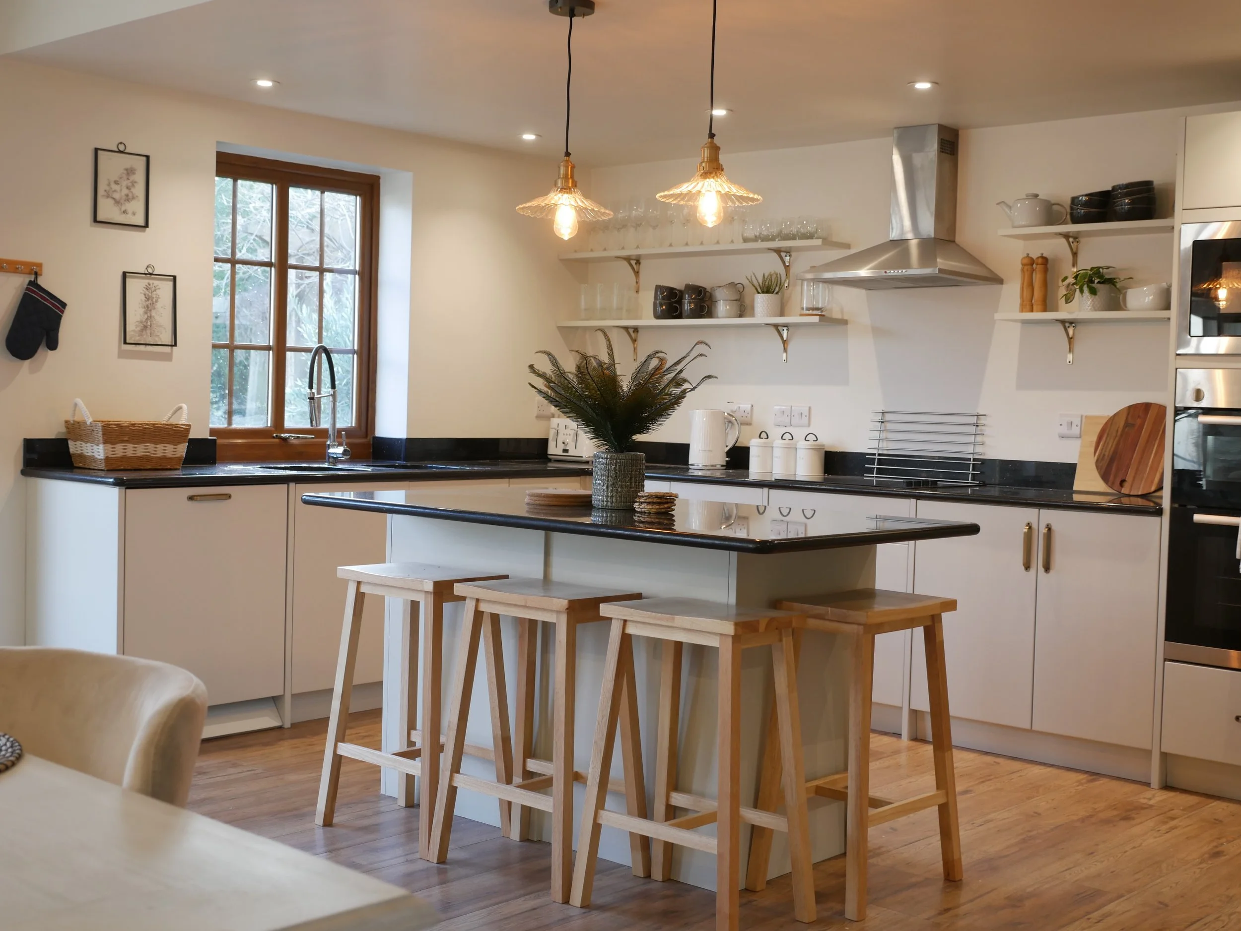 A modern, stylish kitchen featuring crisp white cabinetry paired with a sleek black countertop. The central island is the focal point, adorned with a lush plant in the middle and flanked by two wooden stools for casual seating. Two pendant lights han