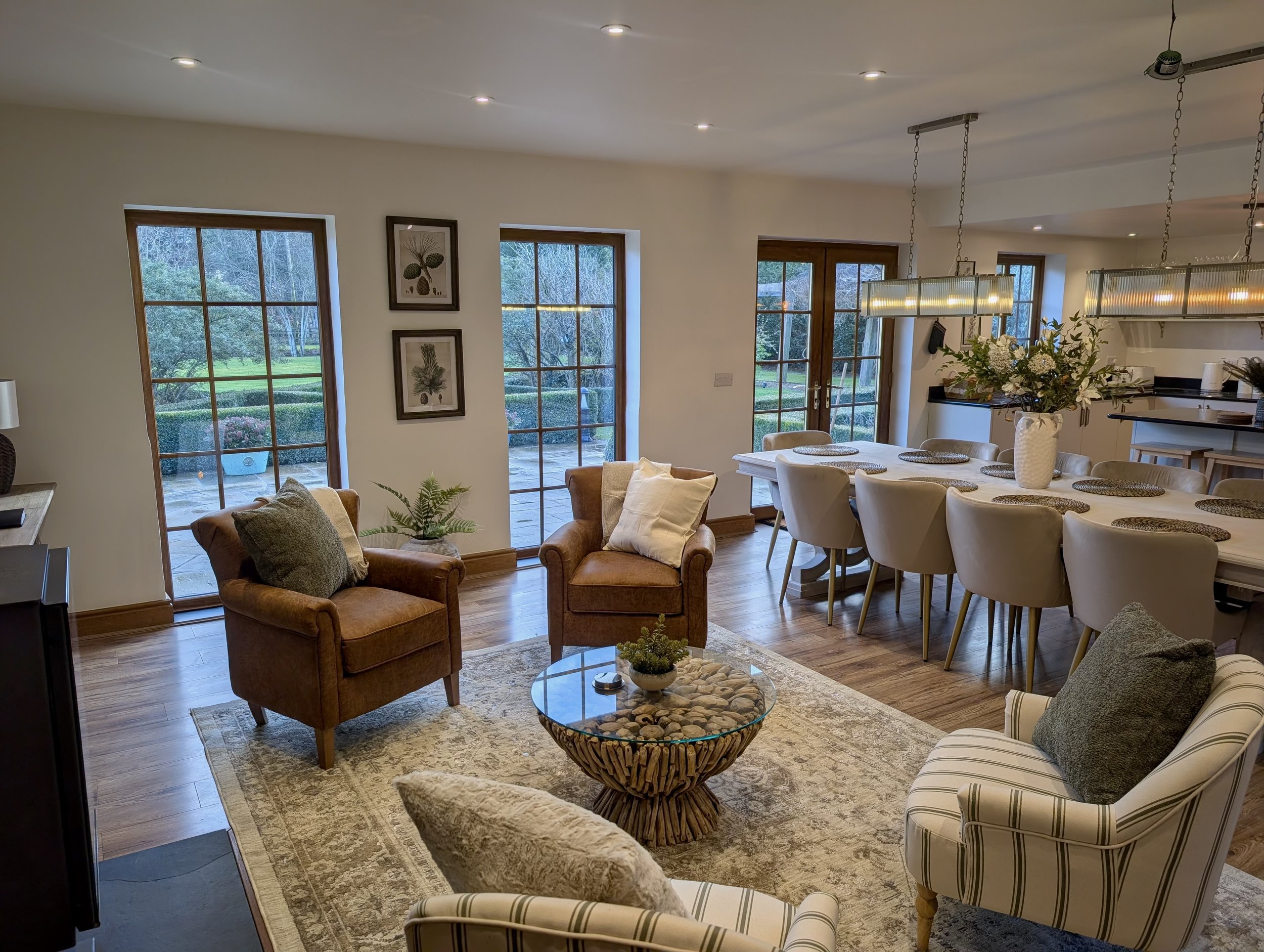 warm contemporary living and dining space with neutral tones and natural textures. Two tan leather armchairs with cushions are positioned in a patterned rug beside a round glass and stone coffee table. Behind them, tall wooden-framed glass doors open
