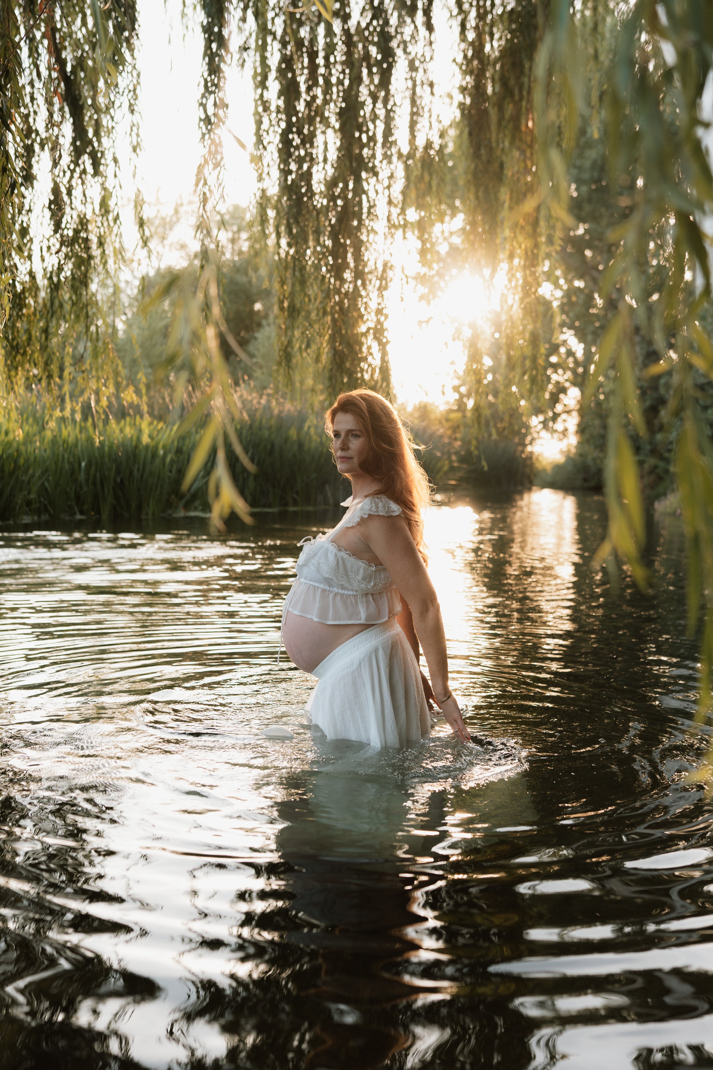 A pregnant woman standing waist-deep in a river beneath willow trees, backlit by warm golden-hour sunlight during an outdoor maternity photography session in Oxfordshire