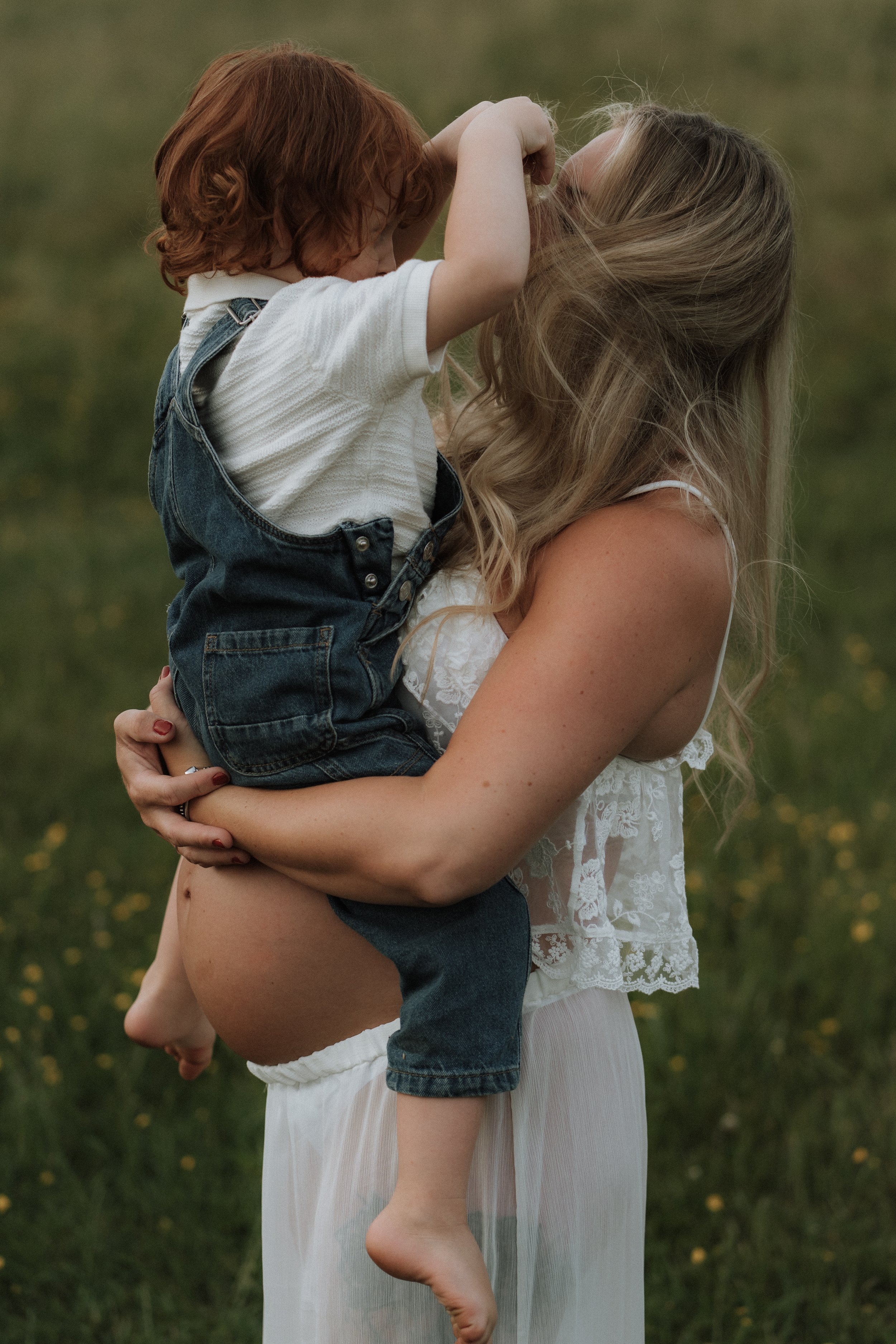 A pregnant woman cuddling her toddler while he plays with her hair during an outdoor maternity photography session in Oxfordshire