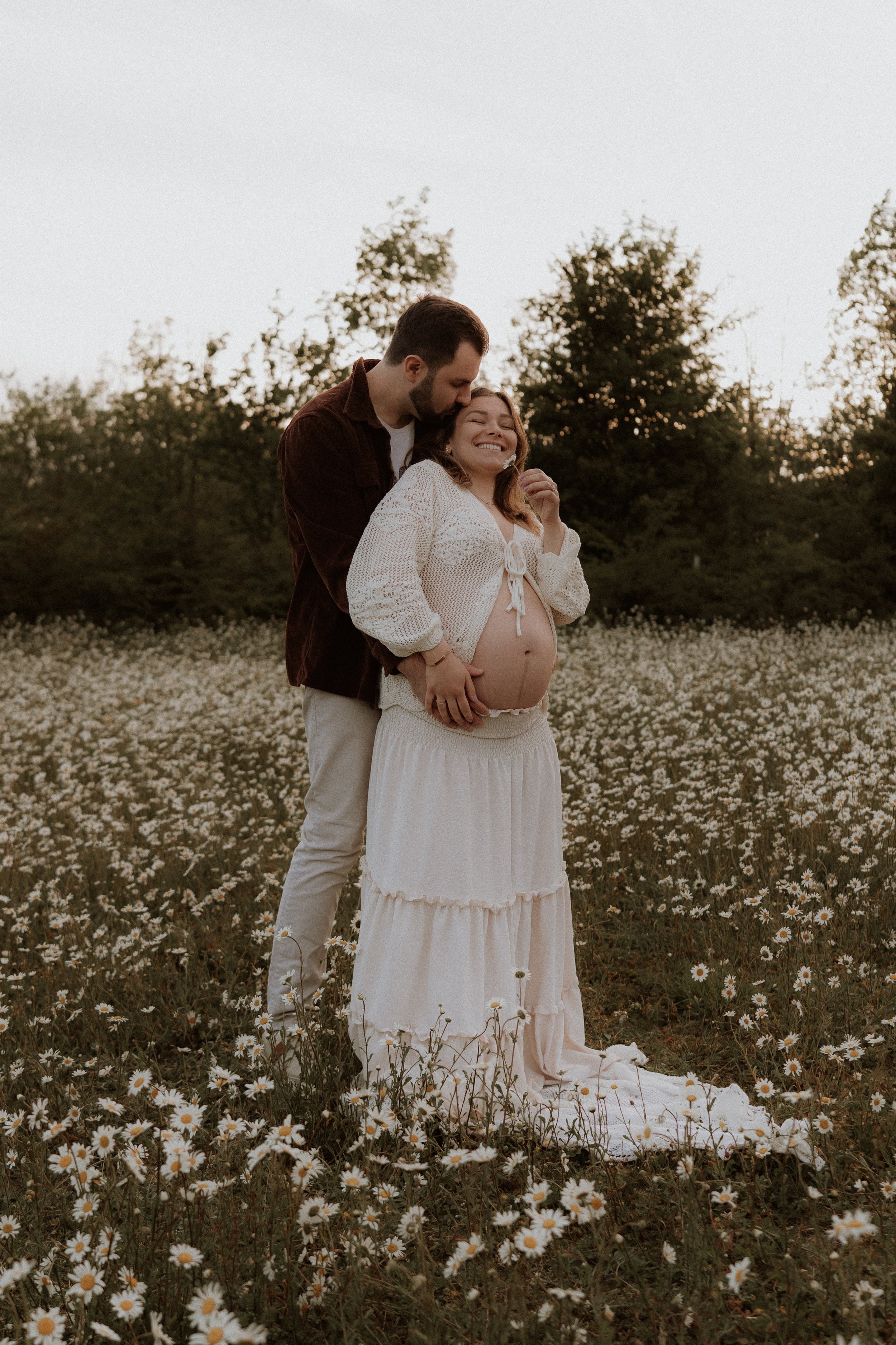 A pregnant woman in a daisy field being embraced by her partner and holding her bump during an outdoor maternity photography session in Oxfordshire