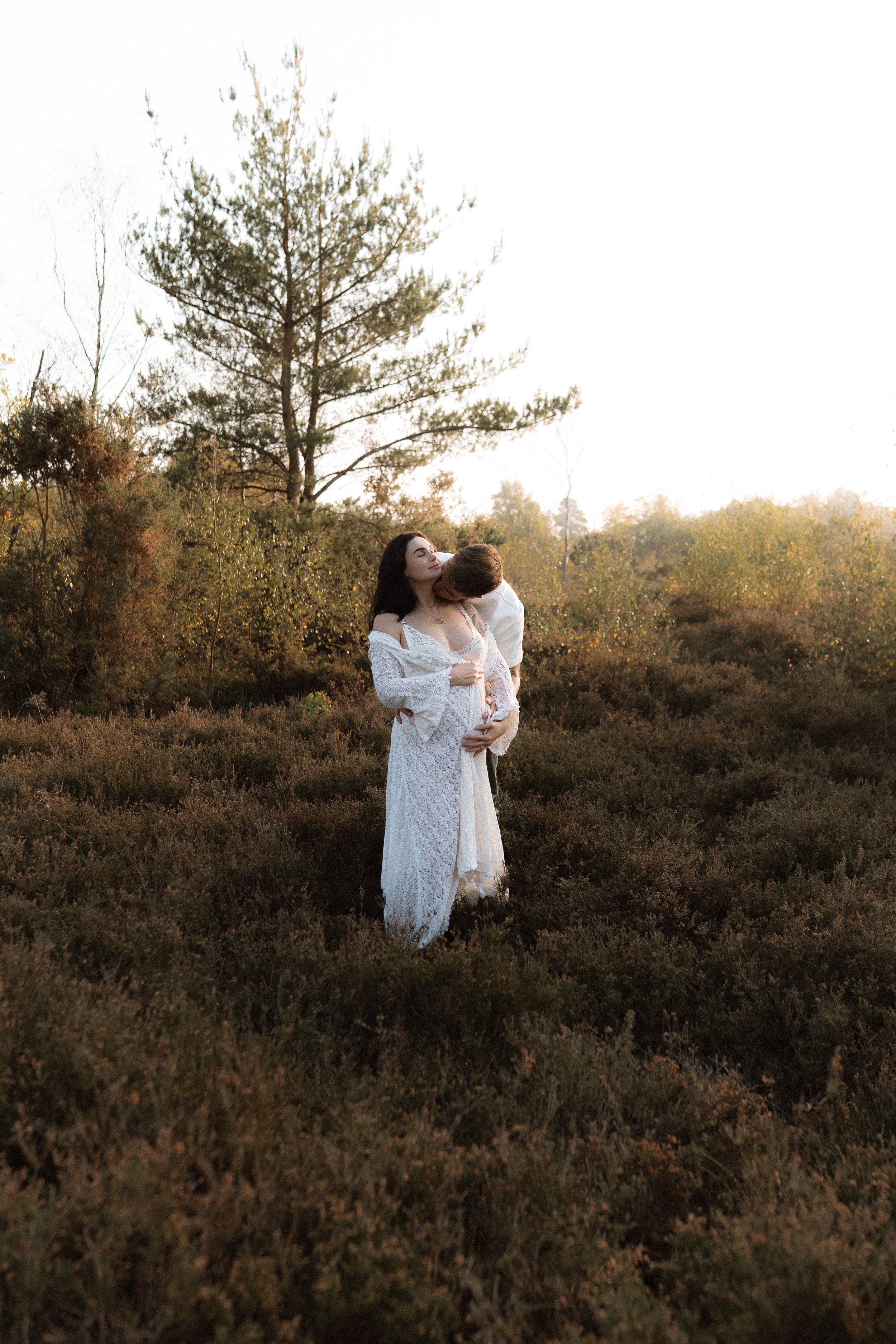 A pregnant woman in a white dress cradling her bump while embracing her partner outdoors among woodland and heather during a maternity photography session in Oxfordshire.