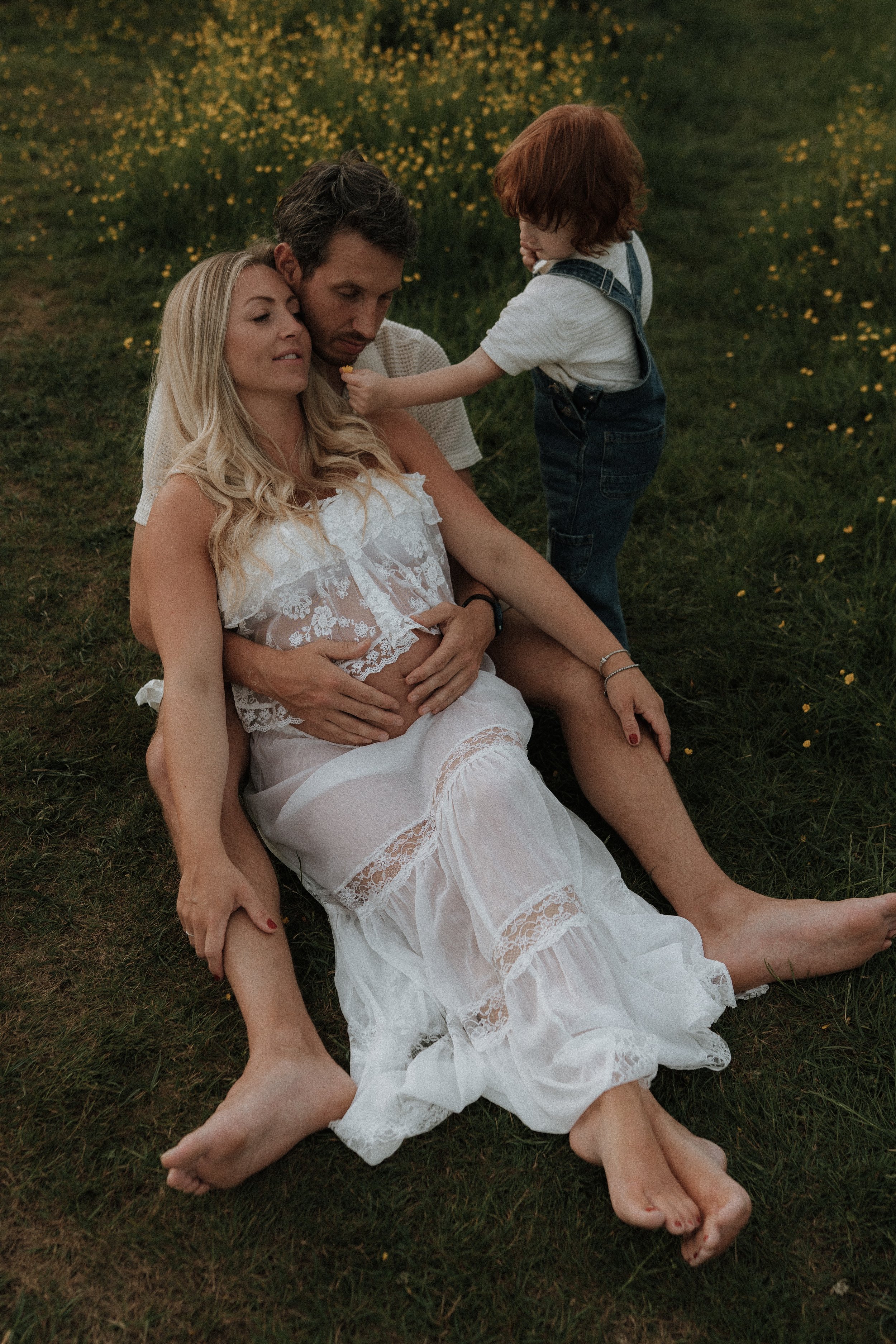 A pregnant woman, lying in her partner's arms as her toddler offers her a flower during an outdoor maternity photography session in Oxford