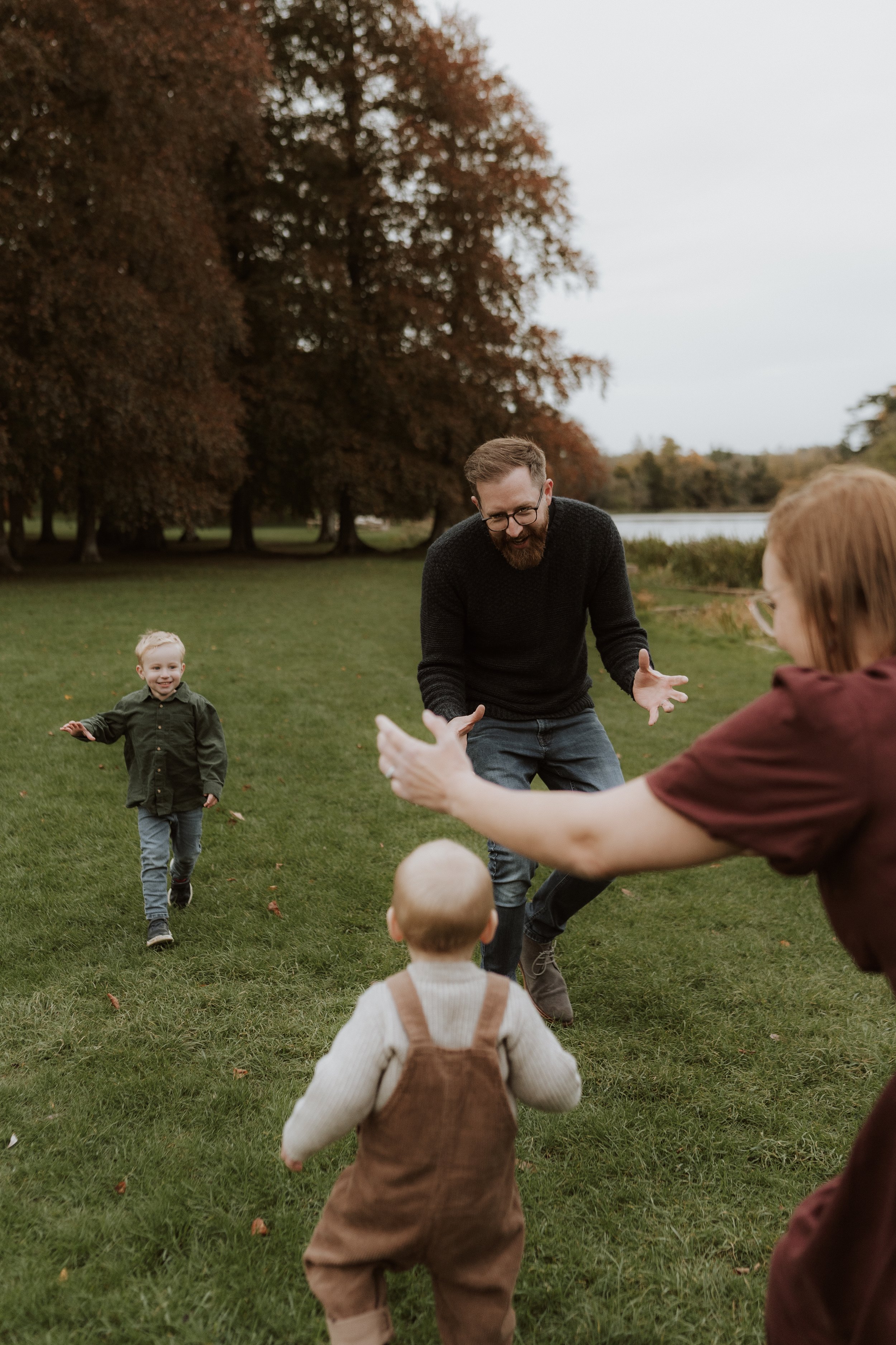 Parents encouraging their toddler’s first steps during an outdoor family photography session in the Cotswolds, captured in natural evening light