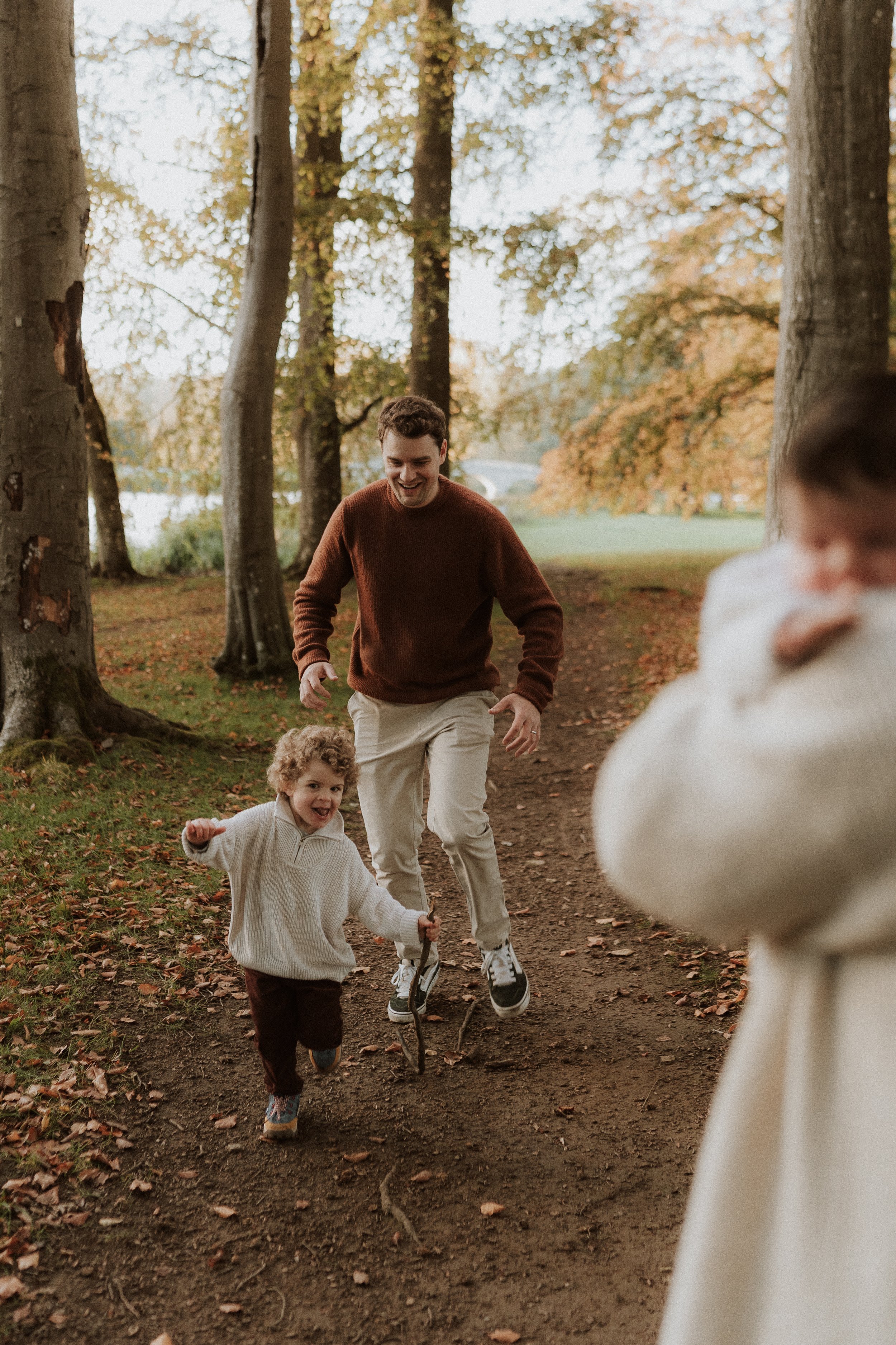 A dad and toddler running through the woods and laughing during an outdoor family photography session in the Cotswolds