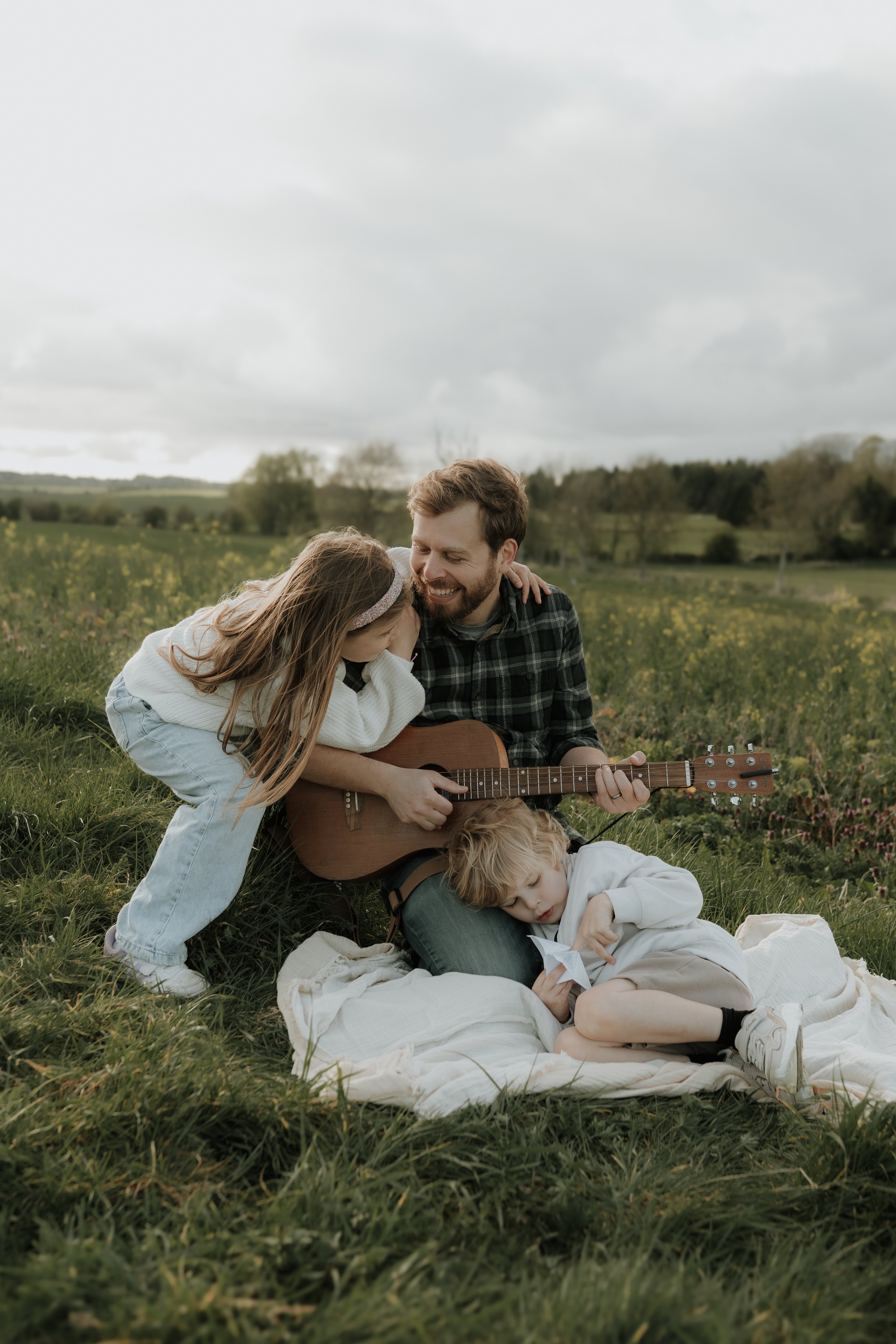 A father playing guitar while cuddling his family during a relaxed outdoor family photography session in the Cotswolds at golden hour.