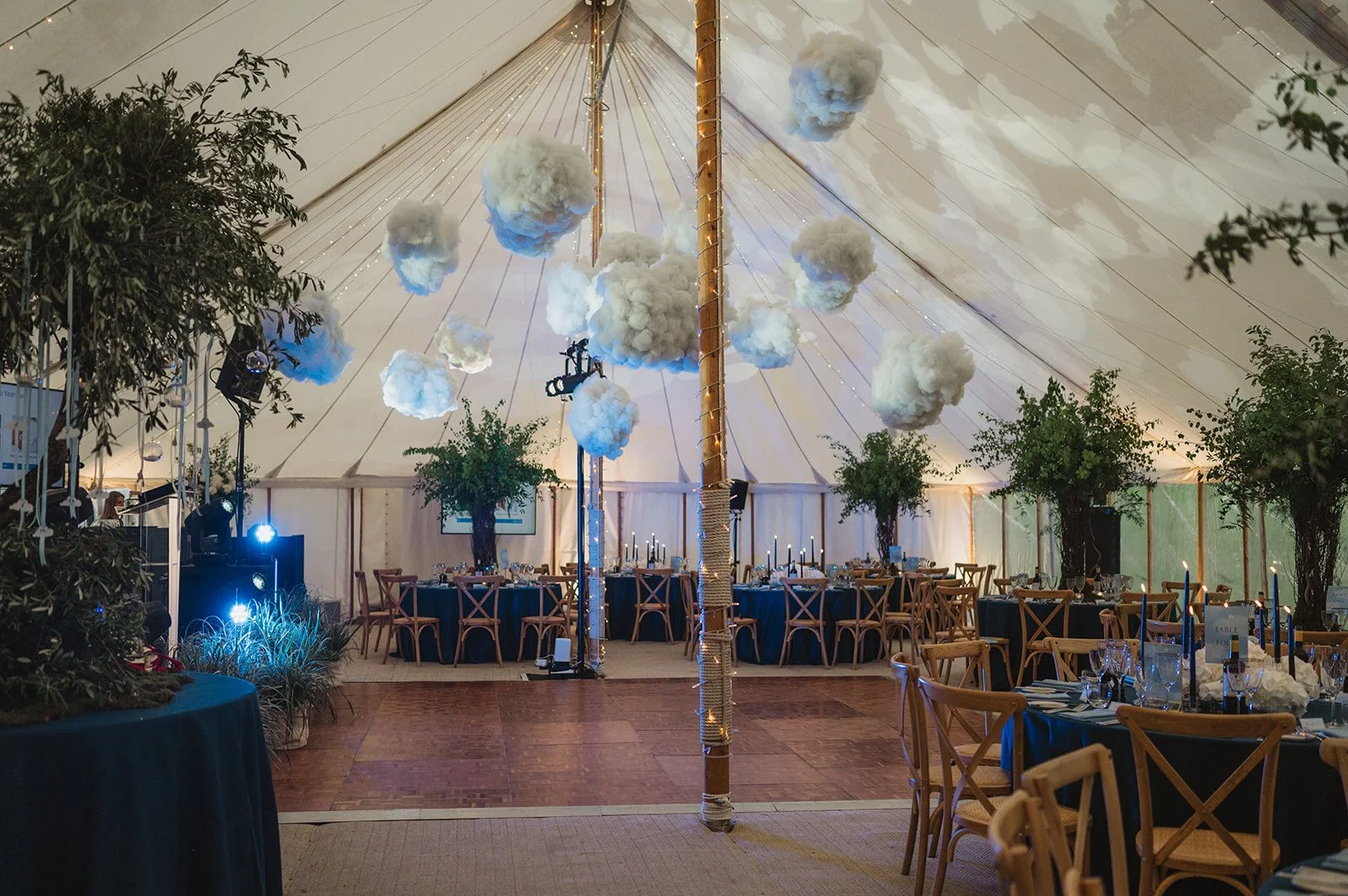 Inside a decorated event tent with cloud-shaped decorations hanging from the ceiling, round tables with blue tablecloths and wooden chairs, some with black candles, and string lights along the central pole.