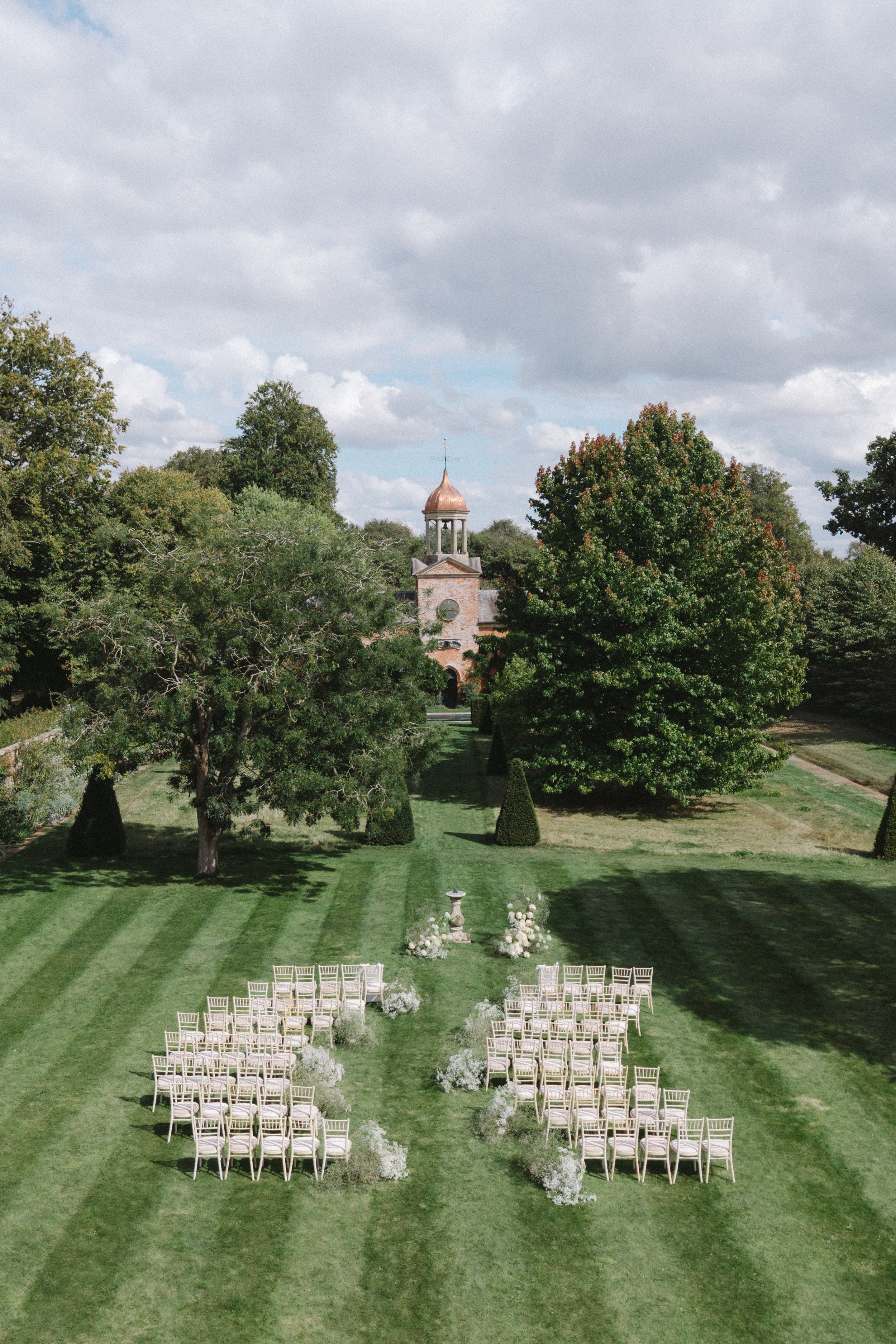 An outdoor wedding ceremony setup on a lush green lawn with rows of white chairs, floral decorations, and a decorative urn, with trees, a brick building with a bell tower, and a partly cloudy sky in the background.