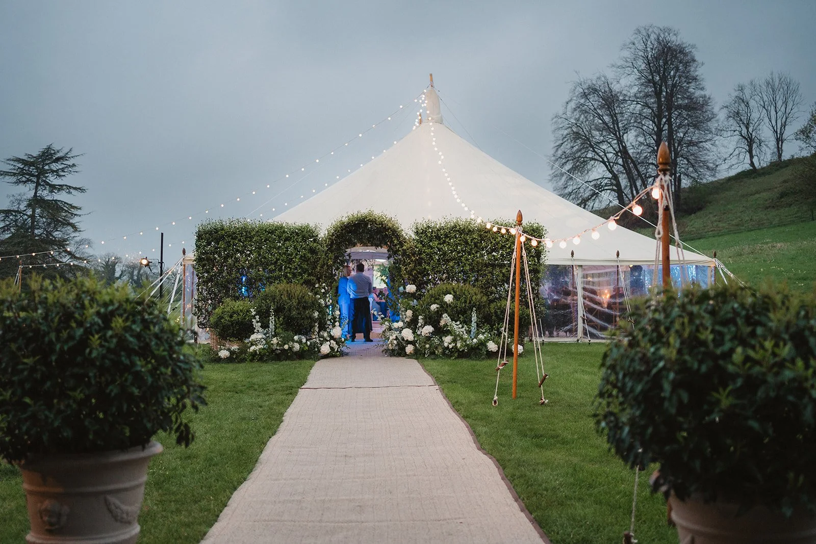 A large white event tent with string lights is set up outdoors on a grassy area, decorated with flowers and surrounded by trees in the evening.