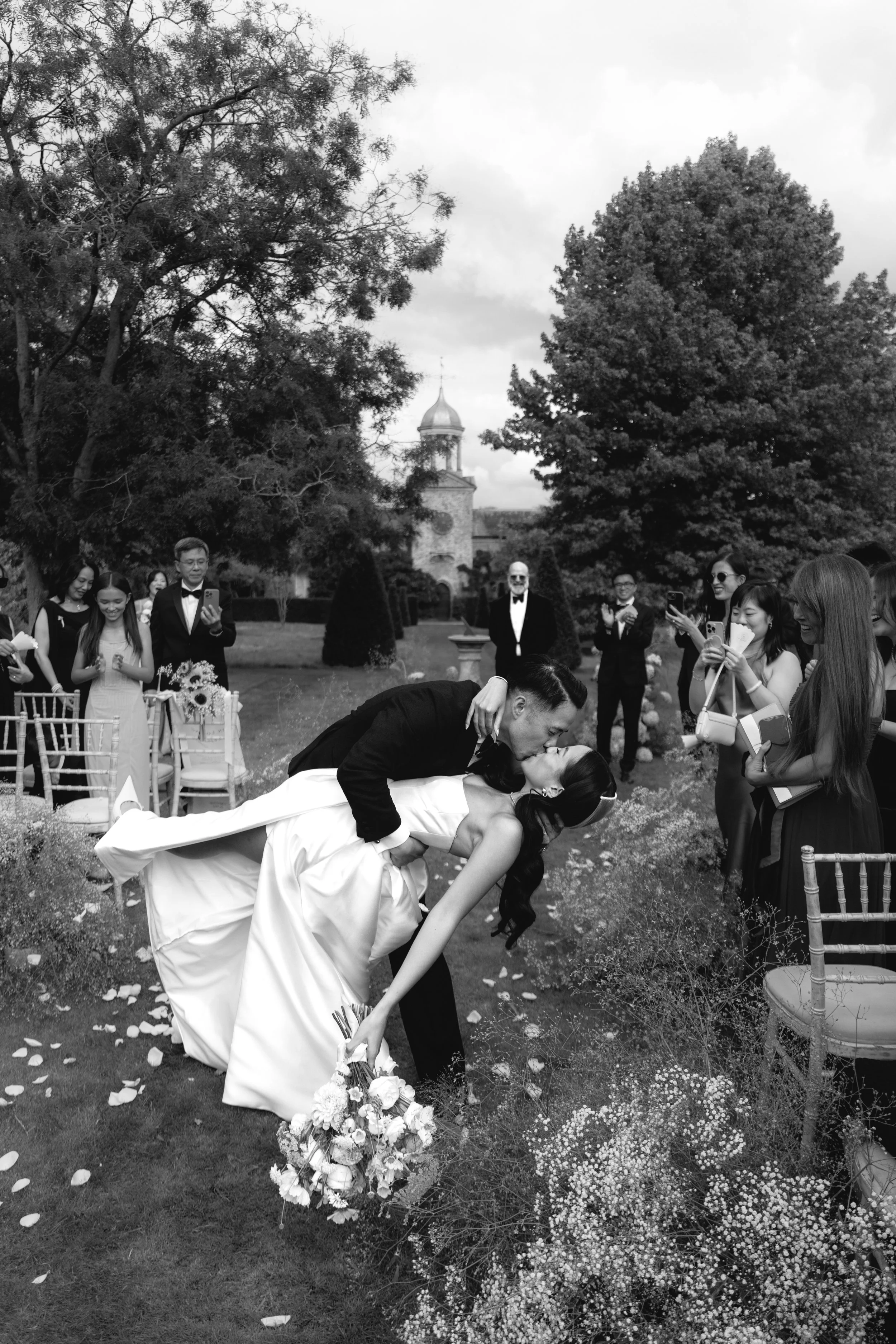 A black and white photo of a wedding celebration outdoors. The bride and groom are kissing as the groom dips the bride. The bride is holding a bouquet of flowers. Wedding guests surround them, some taking photos. The setting includes trees, a grassy 