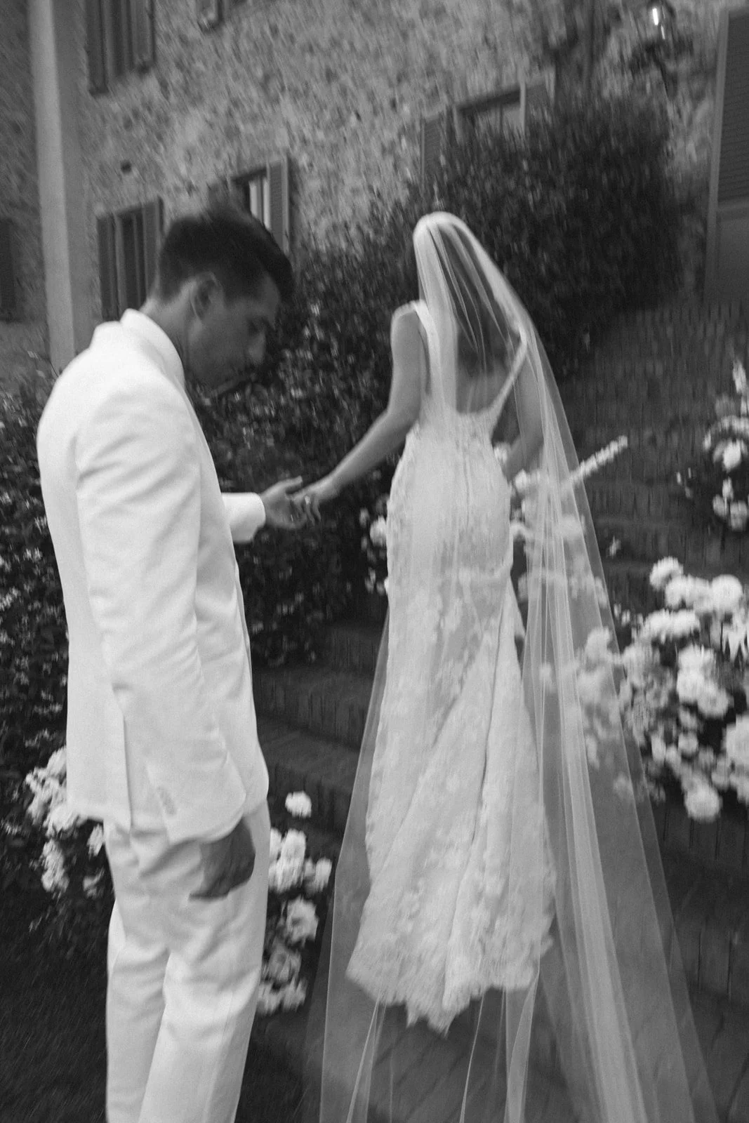 A groom in a white suit gently holding the hand of a bride in a lace wedding dress and veil during a wedding ceremony outdoors at night, with flowers and a brick wall in the background.