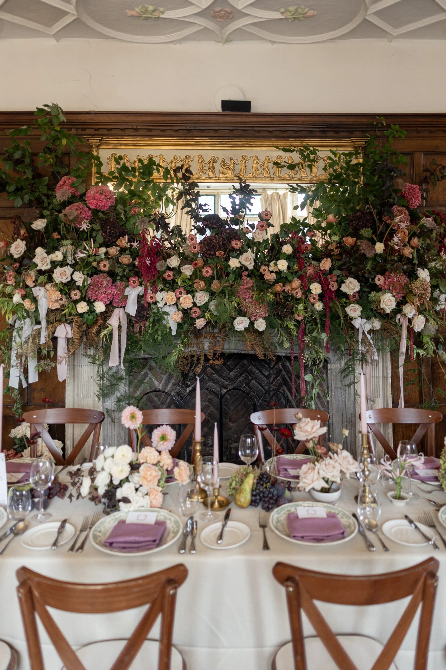Elegant dining table set for a celebration with a floral centerpiece, surrounded by chairs, with a decorated fireplace and floral arrangements in the background.