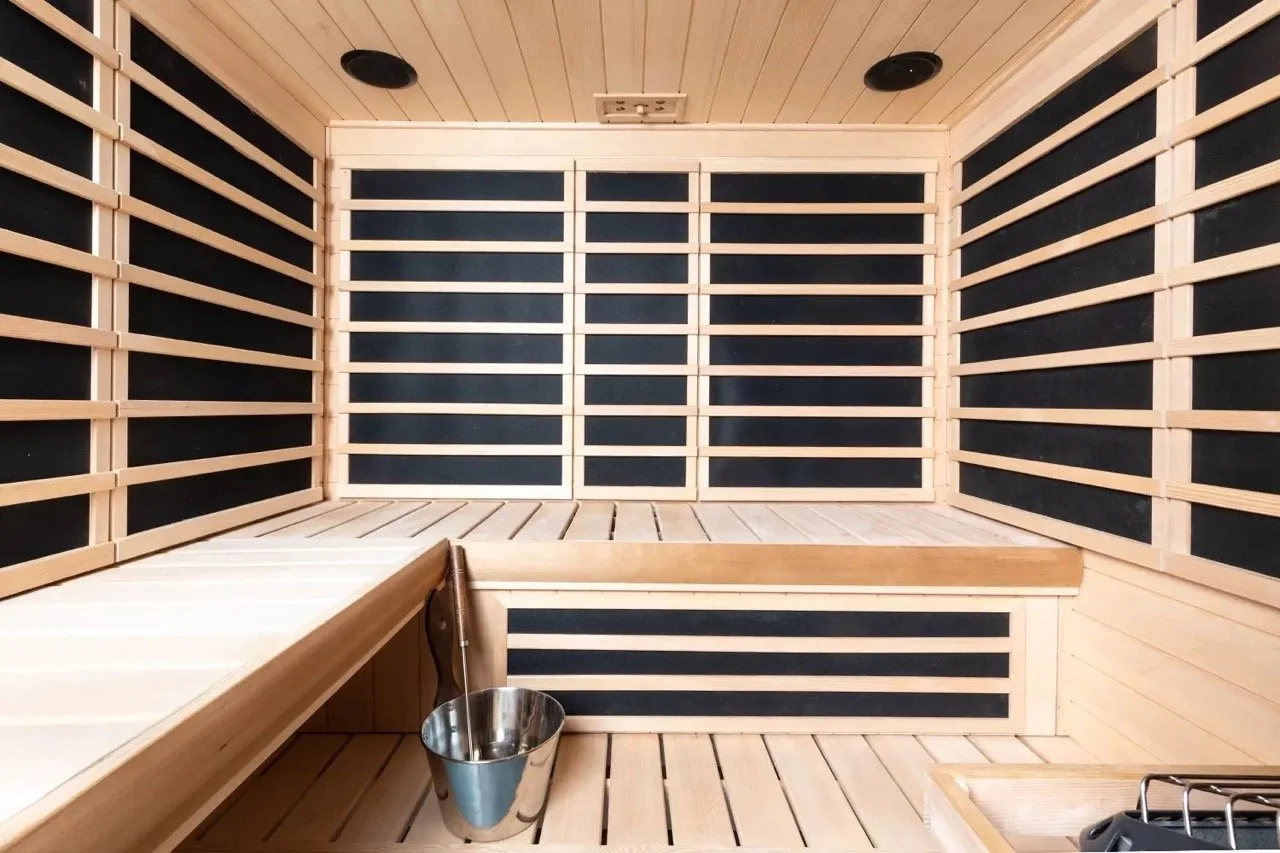 Interior of a modern infrared sauna with wooden benches and walls, featuring black heating panels and a metal bucket on the floor.