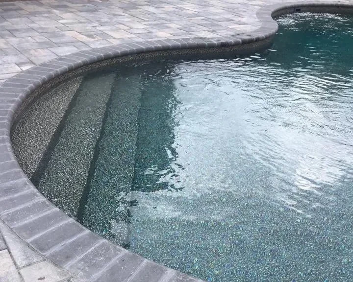 A close-up view of a swimming pool with steps leading into the water, surrounded by a gray brick patio.