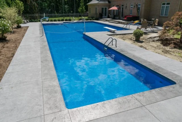 A backyard with a rectangular swimming pool with blue water, surrounded by a concrete deck, next to a house with outdoor furniture and a patio umbrella.