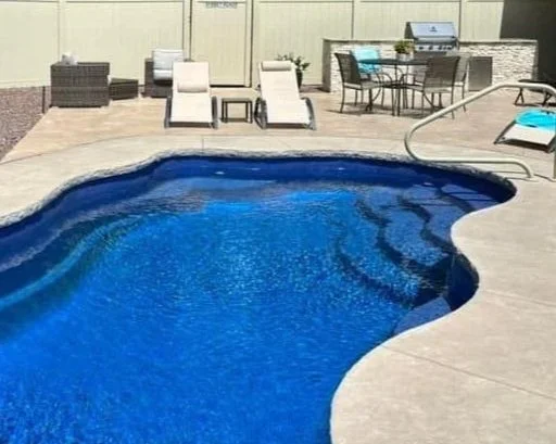 Backyard pool area with two white lounge chairs, a dining table with chairs, and a lounge chair, surrounded by a concrete deck.
