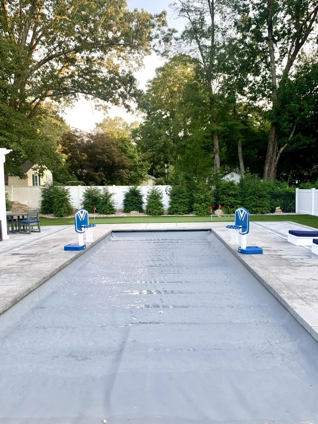 A backyard with a small swimming pool filled with water, two miniature basketball hoops at each end of the pool, surrounded by a patio, green grass, trees, and a white fence.