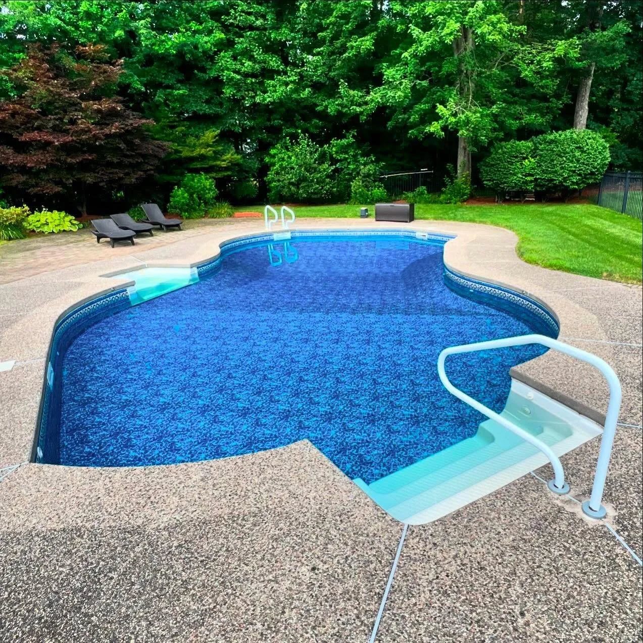 A backyard swimming pool surrounded by grey textured concrete, with three black lounge chairs on the left, greenery in the background, and a black rectangular box on the lawn. The pool has a blue interior with a mosaic pattern and metal handrails at the steps.