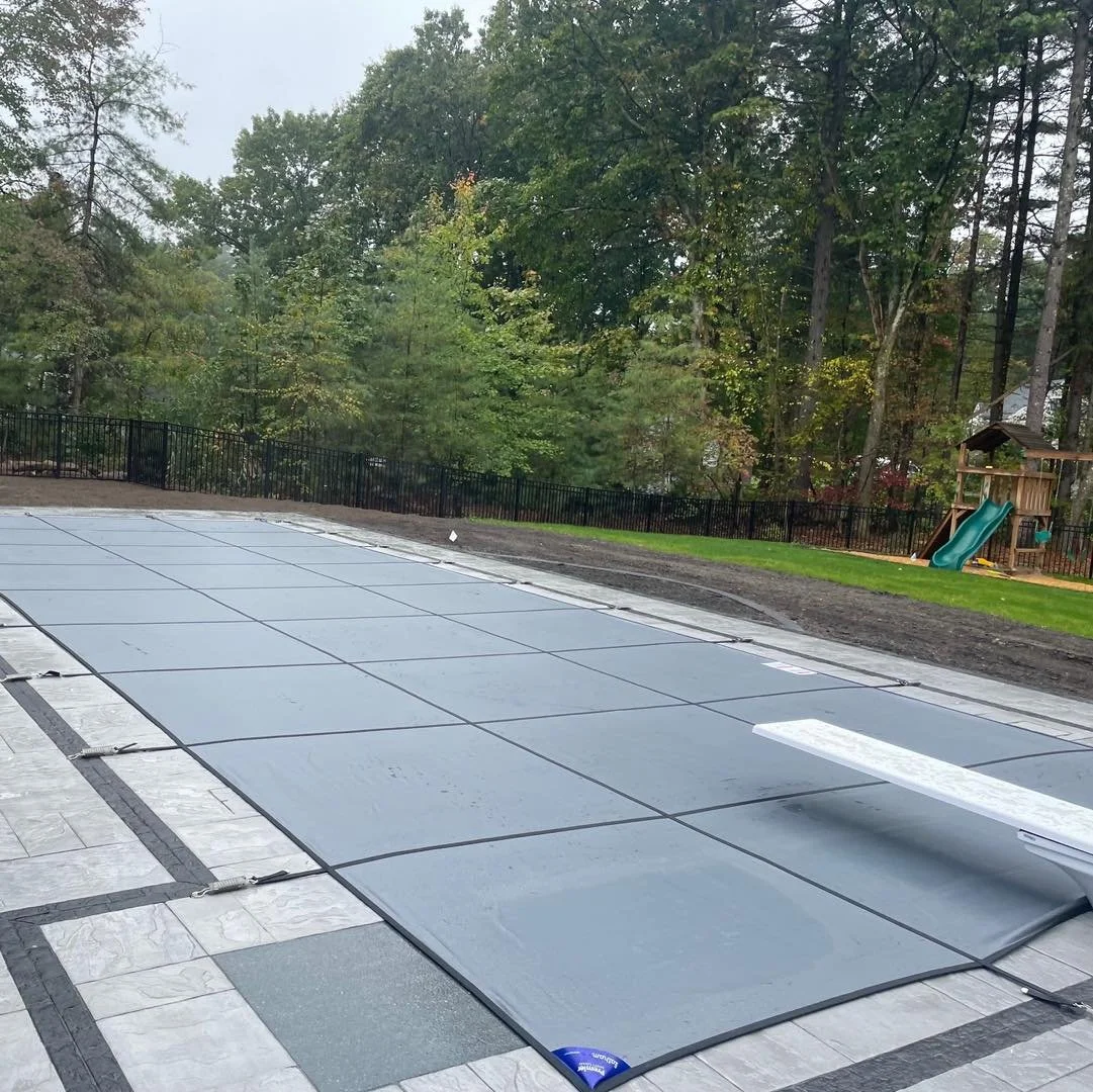 A backyard swimming pool covered with a safety cover, surrounded by a stone patio and grassy yard with a playground in the background, and trees beyond.