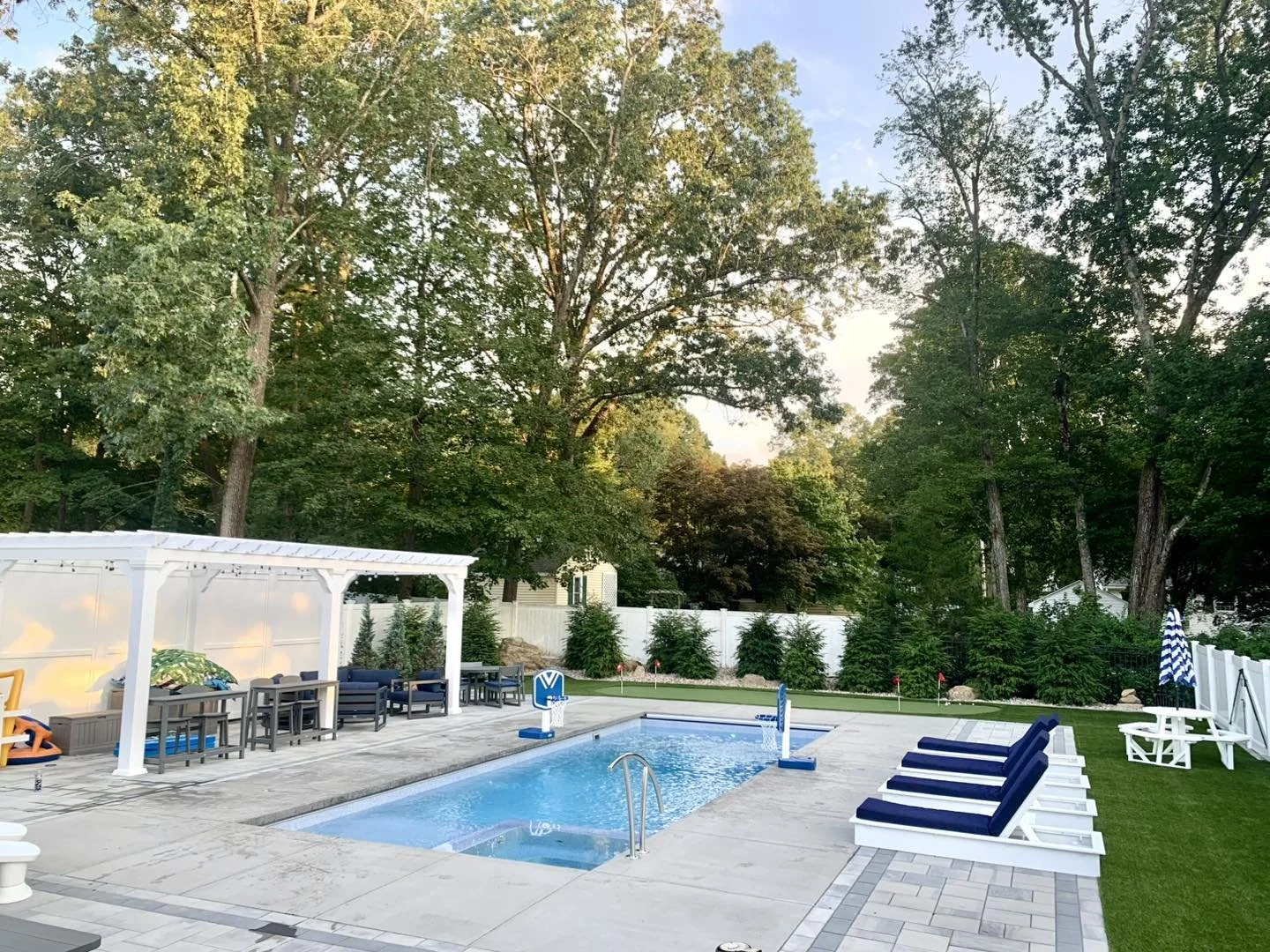 Backyard with a swimming pool, lounge chairs with cushions, and a shaded seating area, surrounded by trees and a white fence.