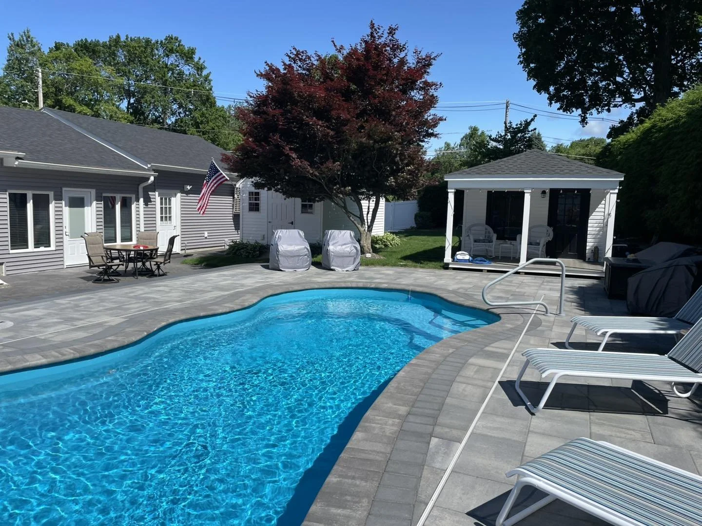 Backyard with a swimming pool, patio furniture, a small white gazebo, a tree with red leaves, and a house with a gray exterior and American flag.
