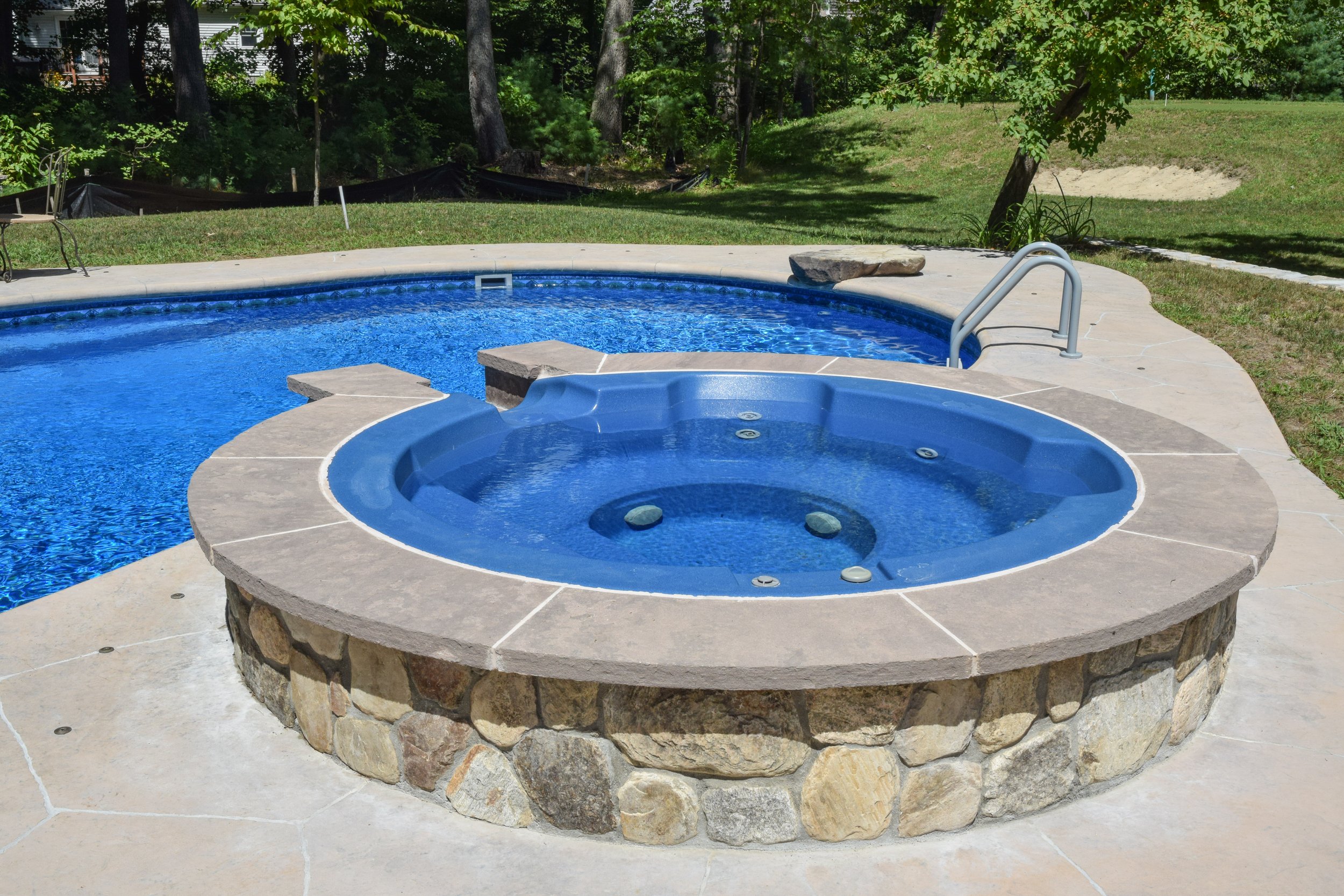 A backyard swimming pool with a built-in hot tub, surrounded by a concrete patio and grassy yard with trees.