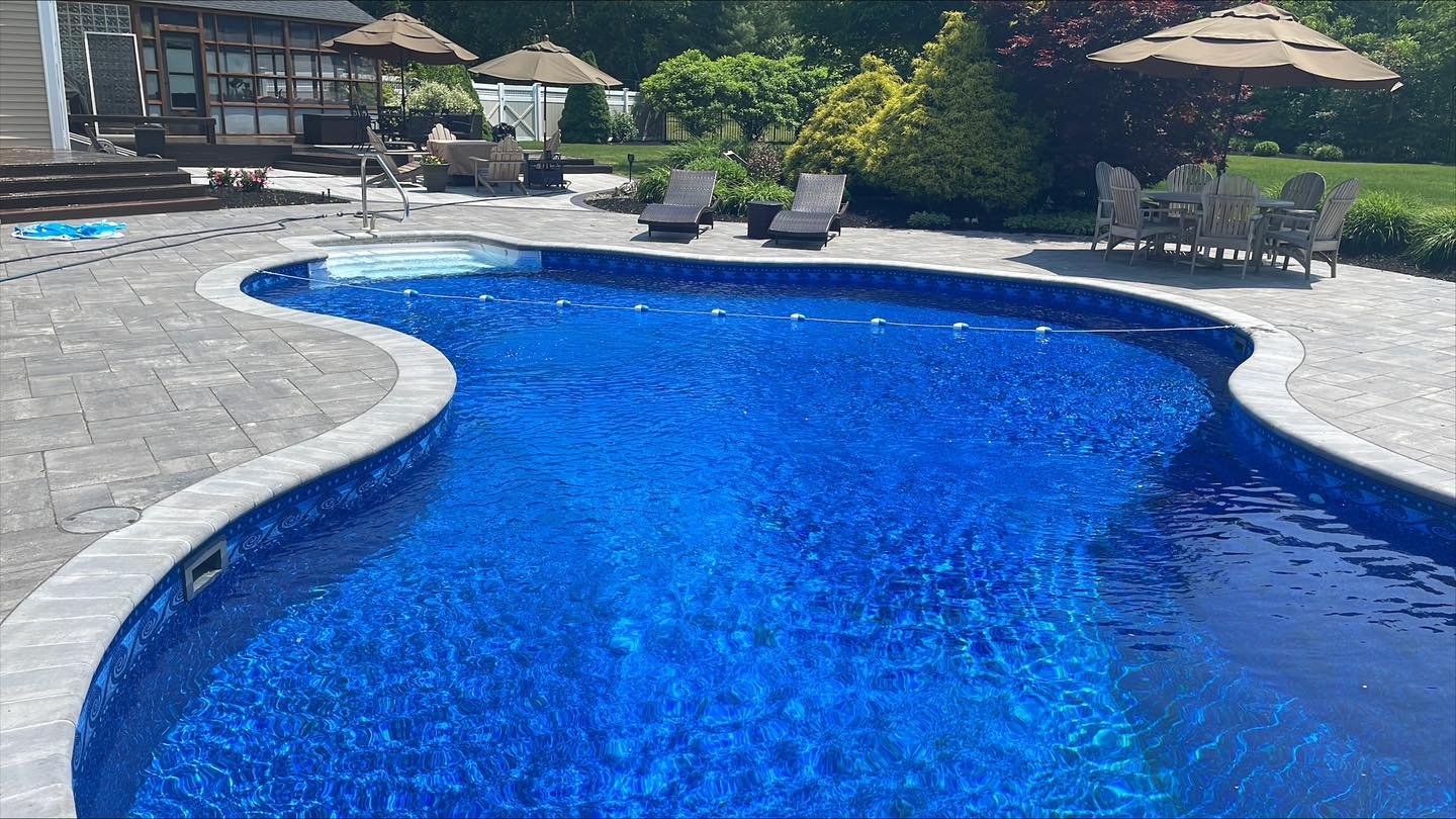 In-ground swimming pool with a hot tub, surrounded by a paved patio area with outdoor furniture and umbrellas, backyard with green grass and trees.