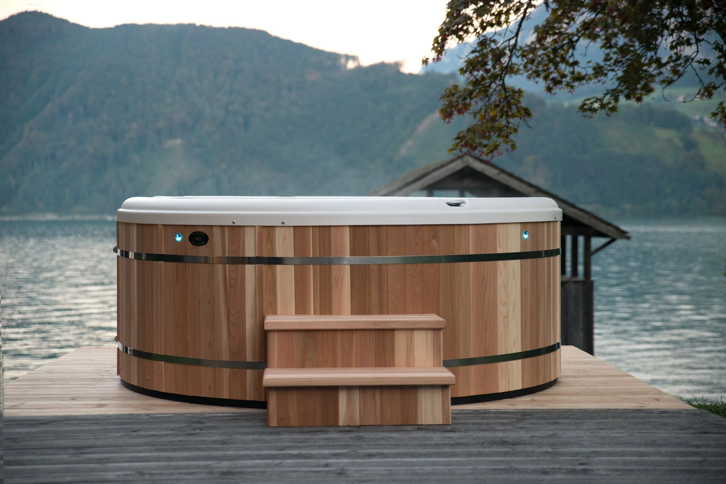 Wood hot tub with stairs on a wooden deck overlooking a lake with mountains in the background.