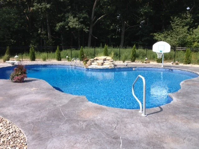 An outdoor backyard swimming pool with a slide, surrounded by greenery and trees, with a basketball hoop nearby.