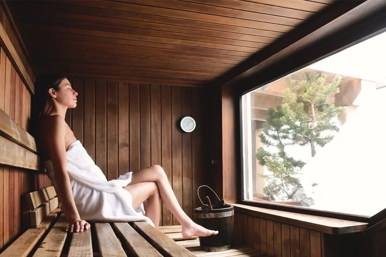 Person relaxing in a wooden sauna, wrapped in a white towel, sitting on a bench near a window with a view of snowy trees.