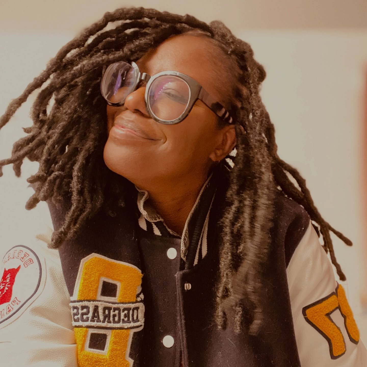 🌻 

ID: A Black woman with freshly retwisted locs smiles and looks upward slightly to the side. She wears oversized round black glasses, an orange sweater, and a varsity jacket with white sleeves and bold yellow letter patches on the front and arm. 