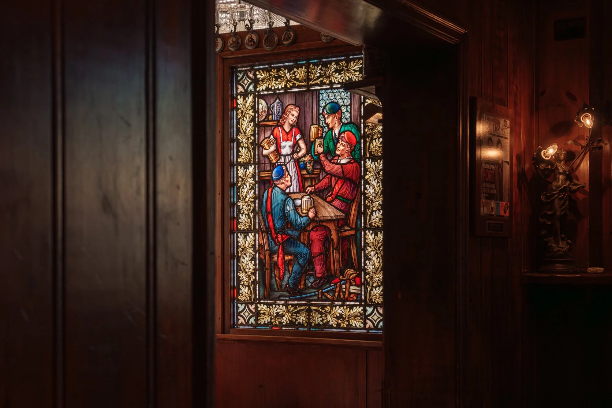 Colorful stained glass window depicting a scene with four people, three seated and one standing, inside a wooden-paneled room decorated with plates and a sculpture.