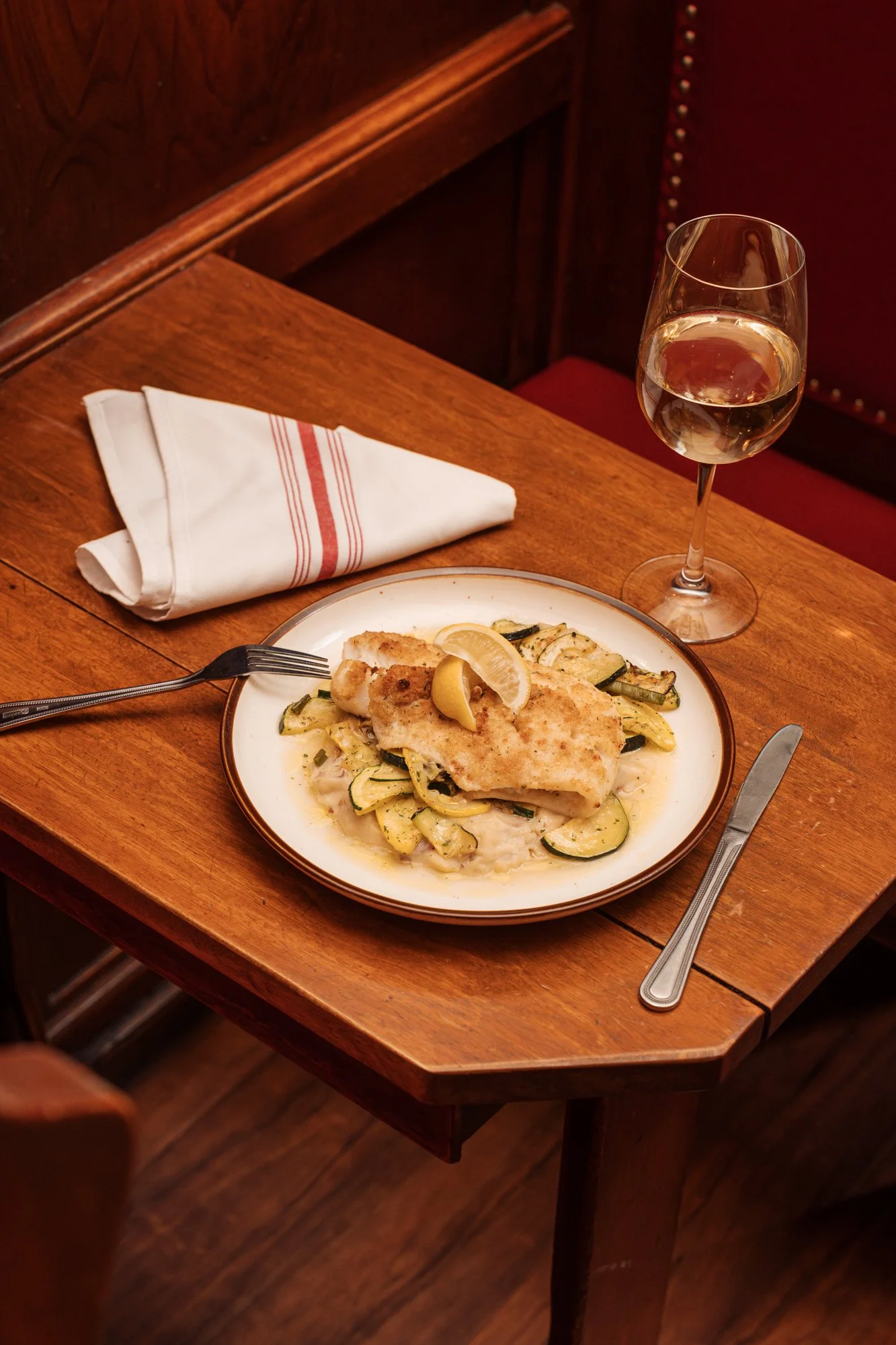 A plate of cooked fish with lemon slices on top, served with zucchini noodles on a wooden table, alongside a glass of white wine and a folded white napkin.