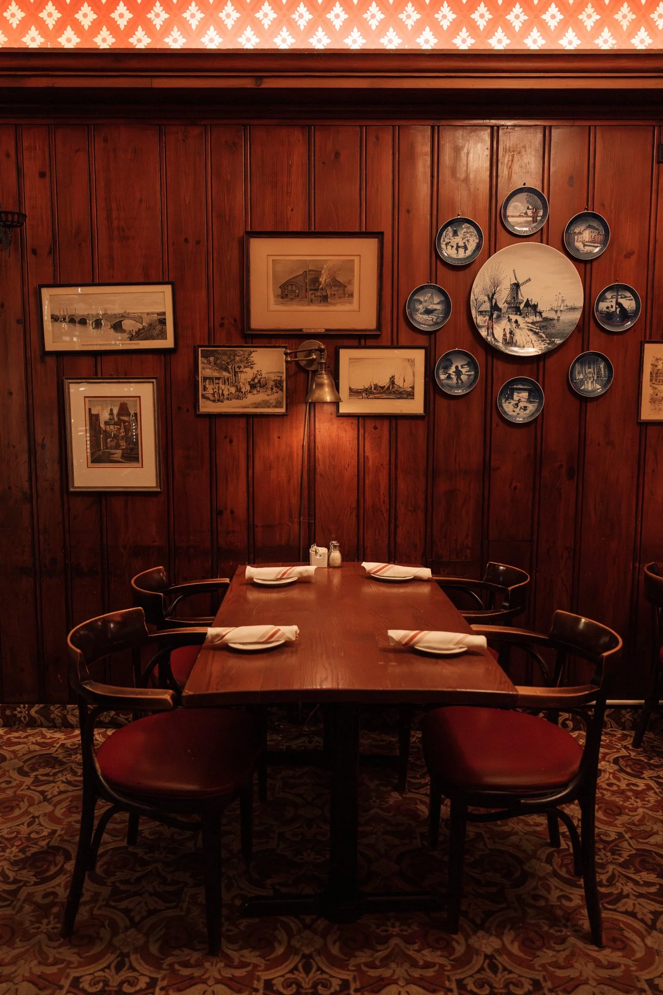 A cozy restaurant corner with a wooden table set for four, with white napkins, salt and pepper shakers, and a small lamp, against a wood-paneled wall decorated with framed pictures and a decorative plate display, with patterned carpeted floor.