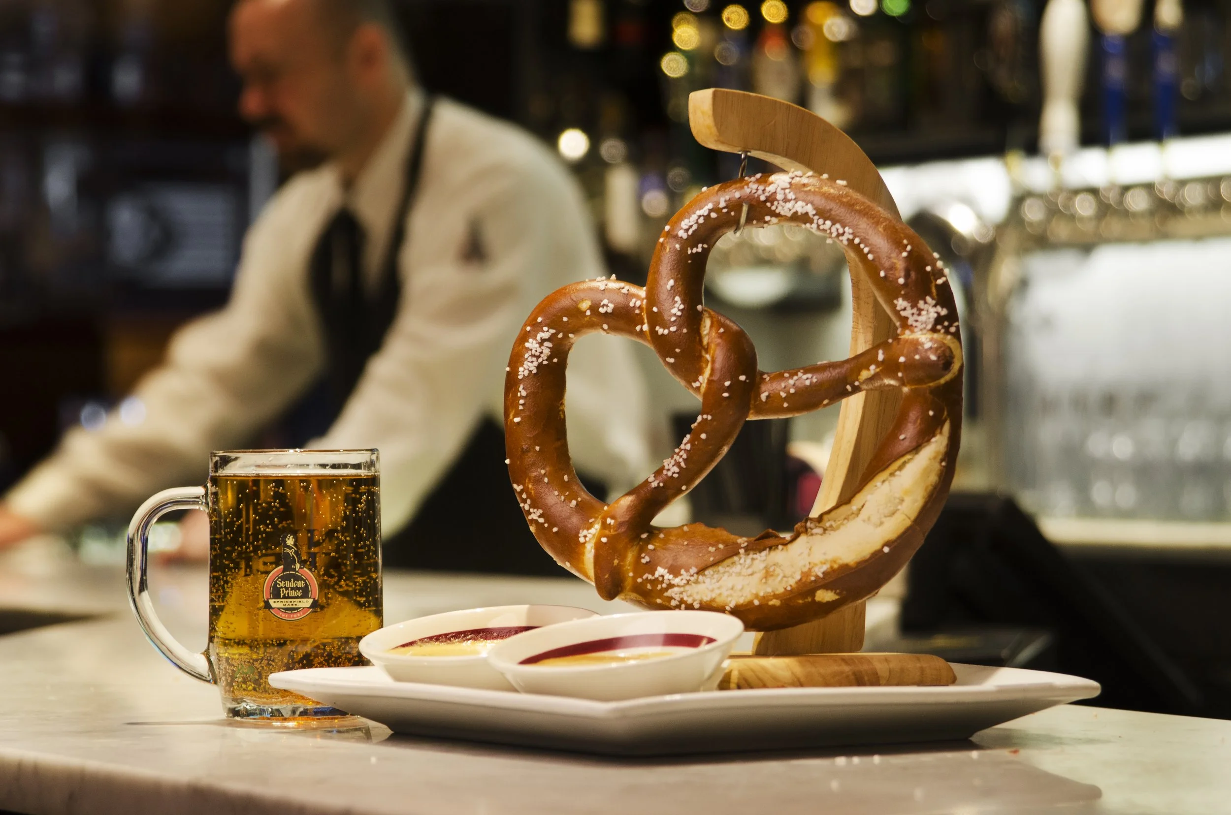 A large, salted pretzel on a wooden stand, with two small bowls of mustard or dip, a mug of beer, and a blurred bartender in the background.