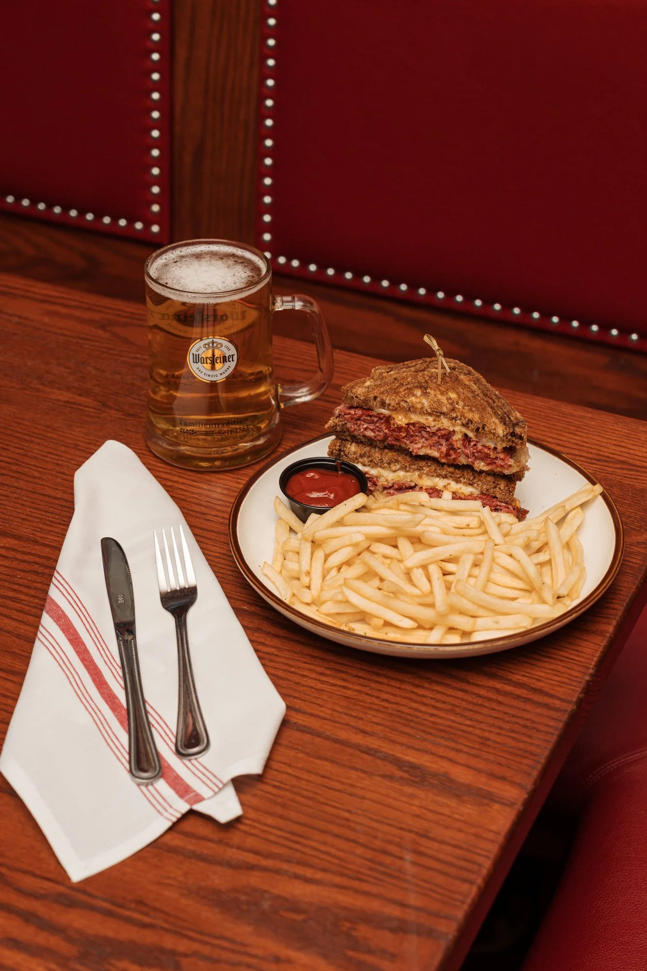 A plate with a grilled sandwich filled with meat and cheese, a side of French fries, and a small cup of ketchup, accompanied by a mug of beer on a wooden table with a white napkin and fork and knife.
