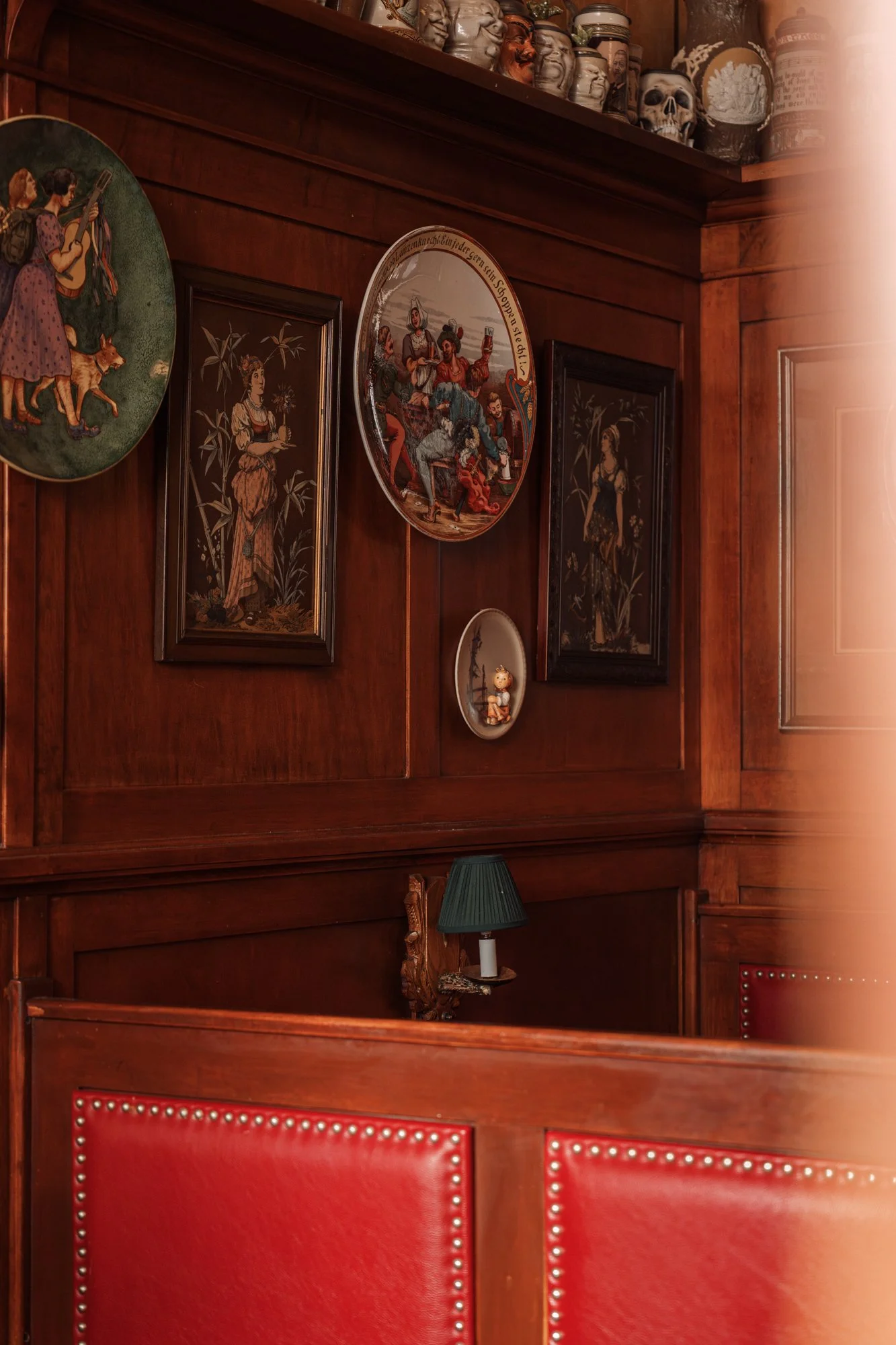 Close-up of a wooden wall decorated with vintage plates and framed artwork, featuring dark wood paneling, a small green lampshade, and a collection of decorative items including skulls and ceramic figures.