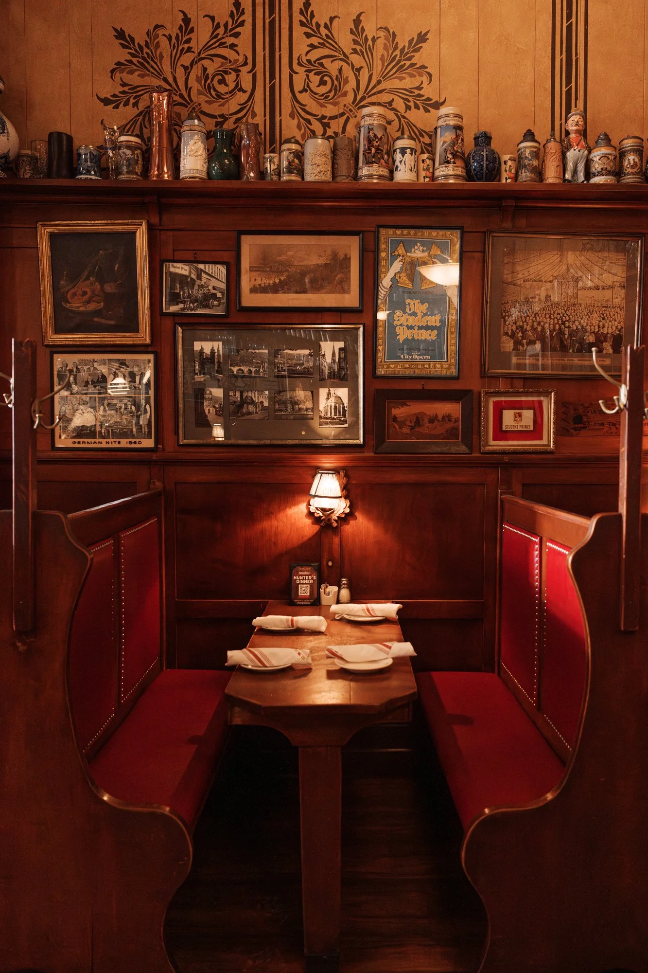 A cozy corner booth in a restaurant with a wooden table set for four, featuring white napkins and plates. The booth has red cushioned seating with wooden accents. The wall above is decorated with a collection of framed photographs and artwork, includ