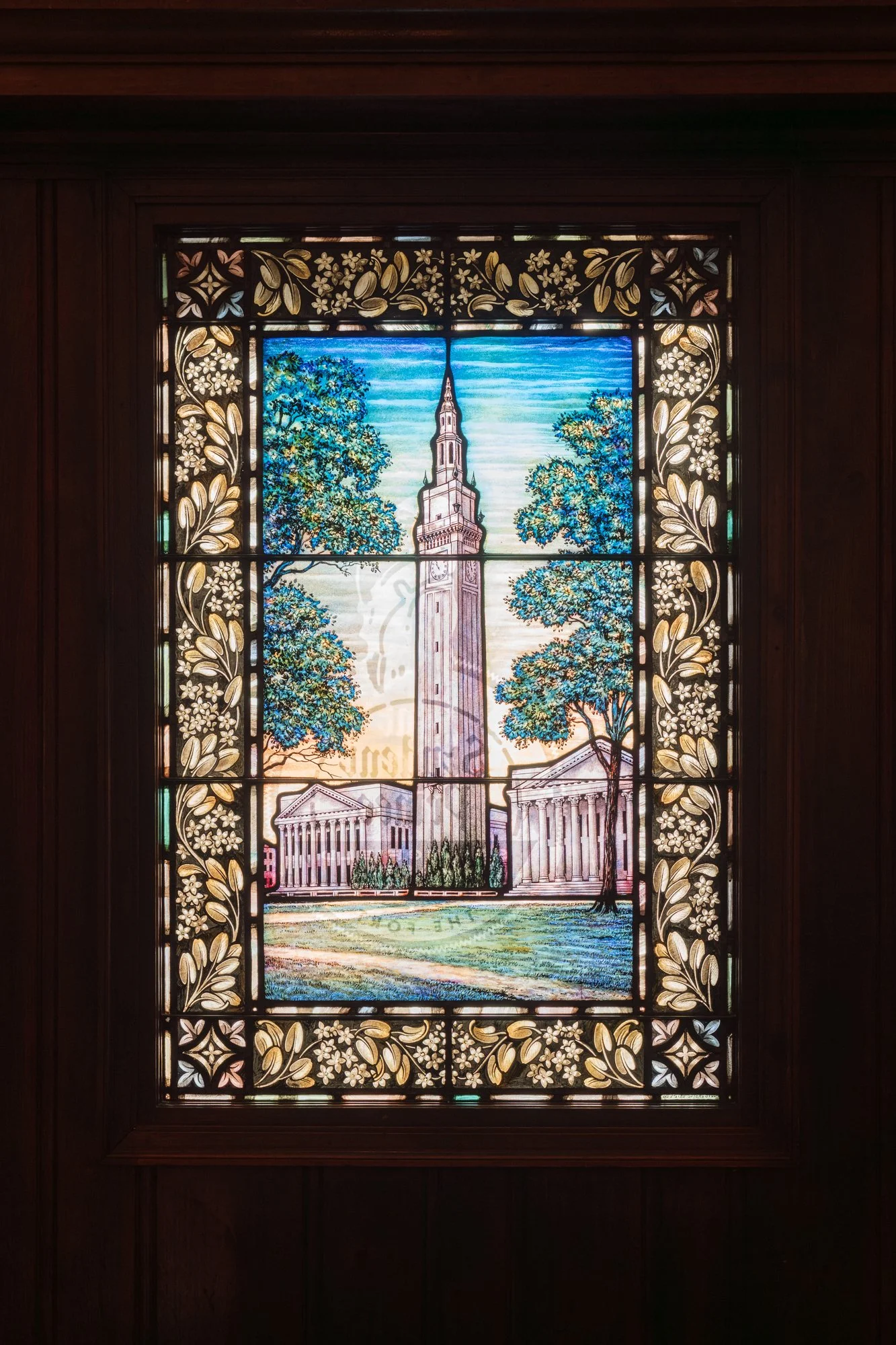 Stained glass window depicting a tall clock tower surrounded by trees and classical buildings with columns, framed by floral and leaf patterns at The Student Prince in Springfield MA