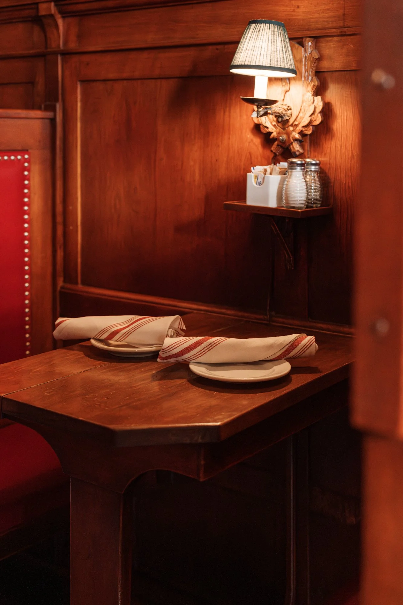 A cozy restaurant corner with a wooden table set with two plates and striped napkins, a wall-mounted lamp, and a shelf with glasses and condiment holders, all featuring warm wooden decor.