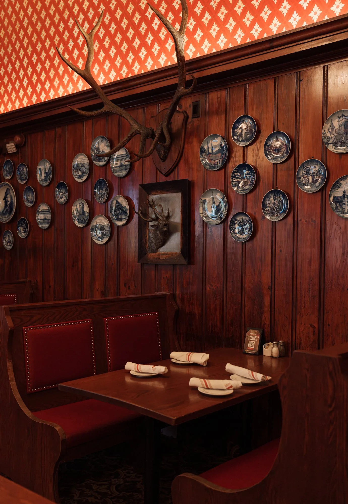 A cozy restaurant corner with wood-paneled walls, red booth seating, neatly folded napkins on a wooden table, and a deer head mount with antlers on the wall. The wall is decorated with numerous blue and white plates and a mounted deer head with antle