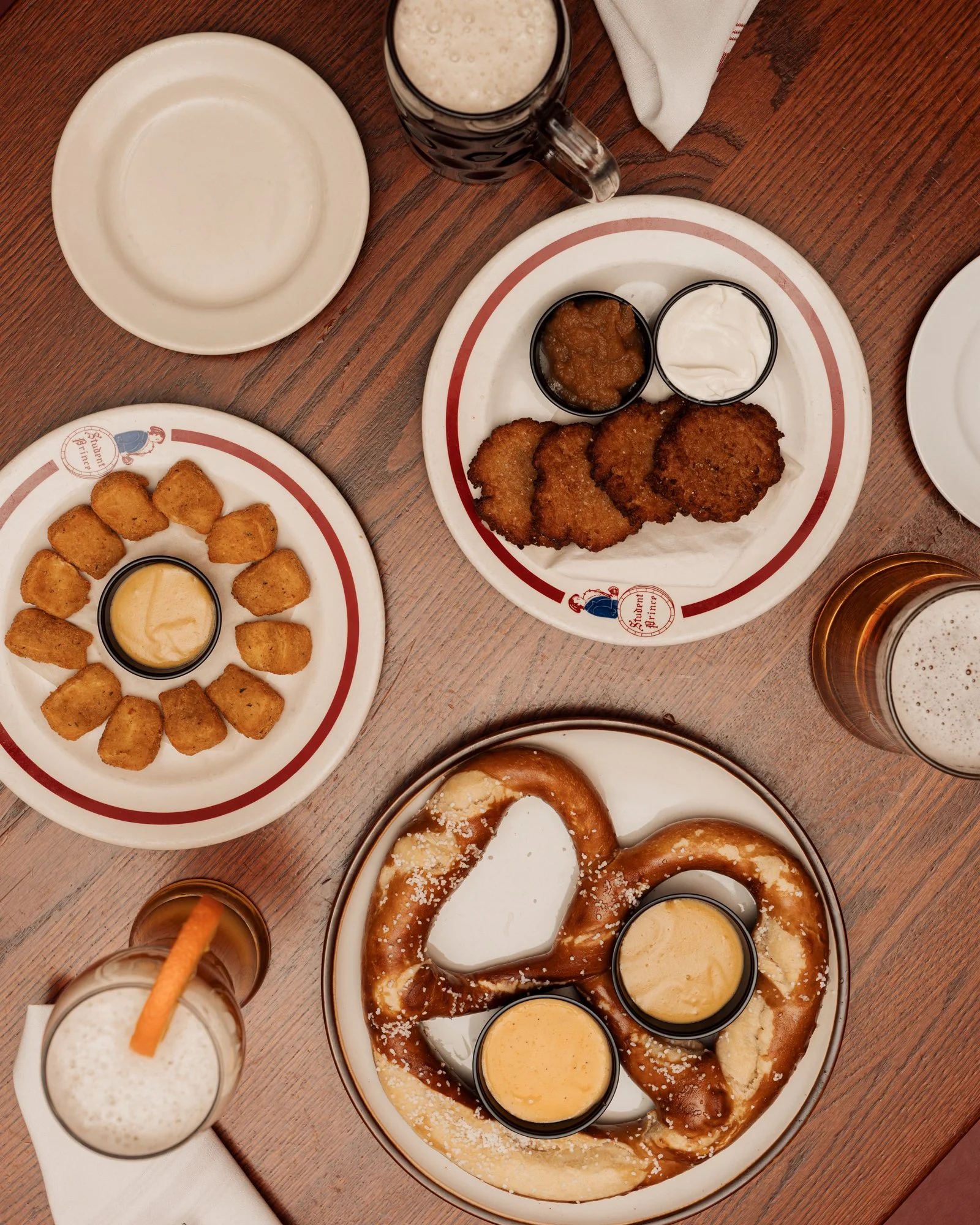 Overhead view of a wooden table with plates of food and drinks, including a large pretzel with dipping sauces, fried chicken tenders with sauces, fried cheese nuggets with a dipping sauce, and glasses of beer and a milkshake.