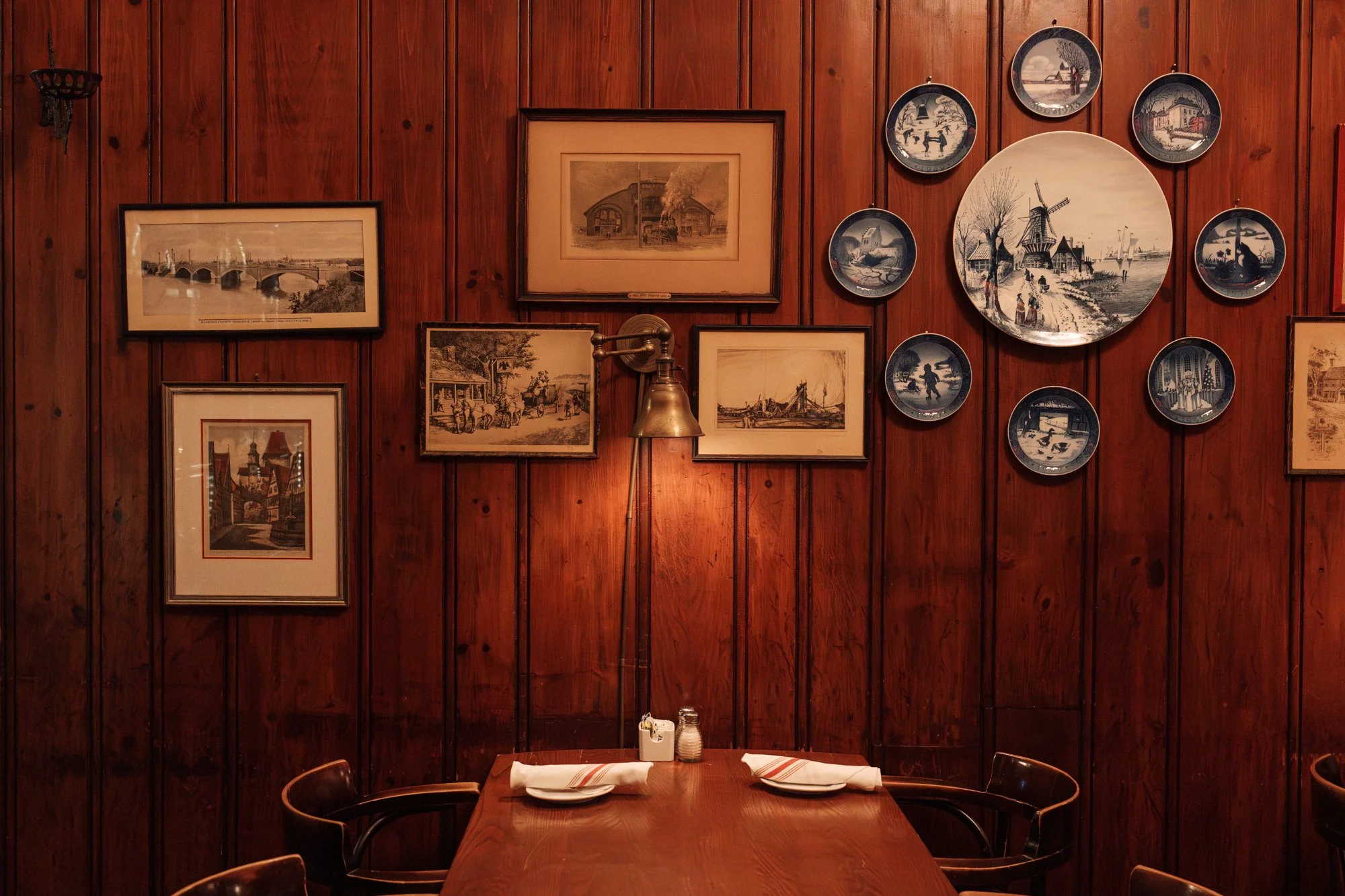 Interior of a restaurant with a wooden-paneled wall decorated with framed vintage artwork and blue and white plates arranged around a central larger plate depicting a windmill scene. A wooden table is set with plates, napkins, and condiments.