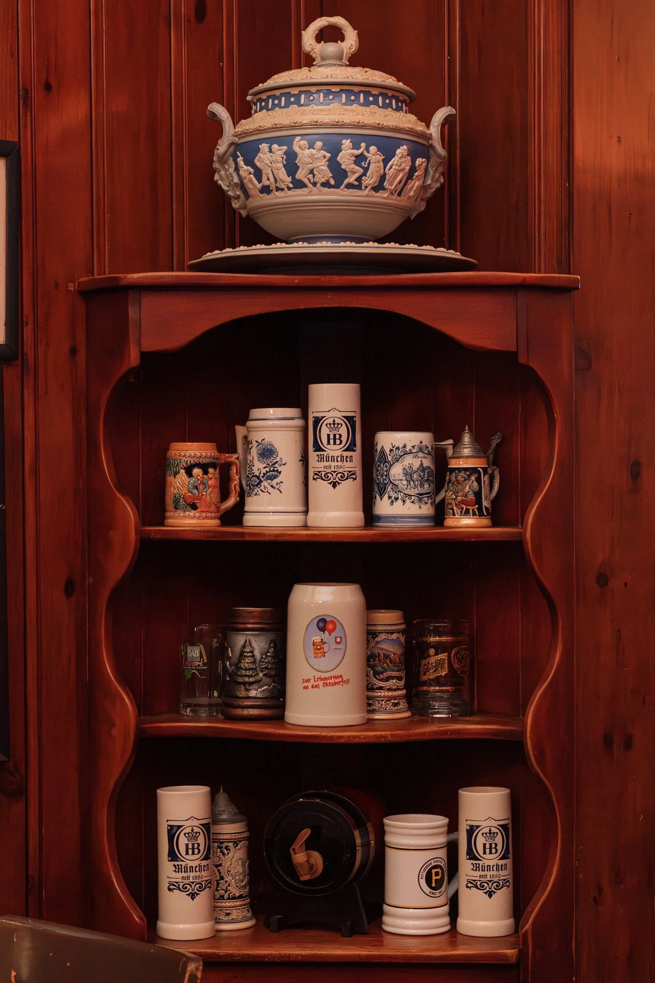 A wooden display cabinet with traditional German beer steins, decorative pottery, and a large ornate tureen on top.