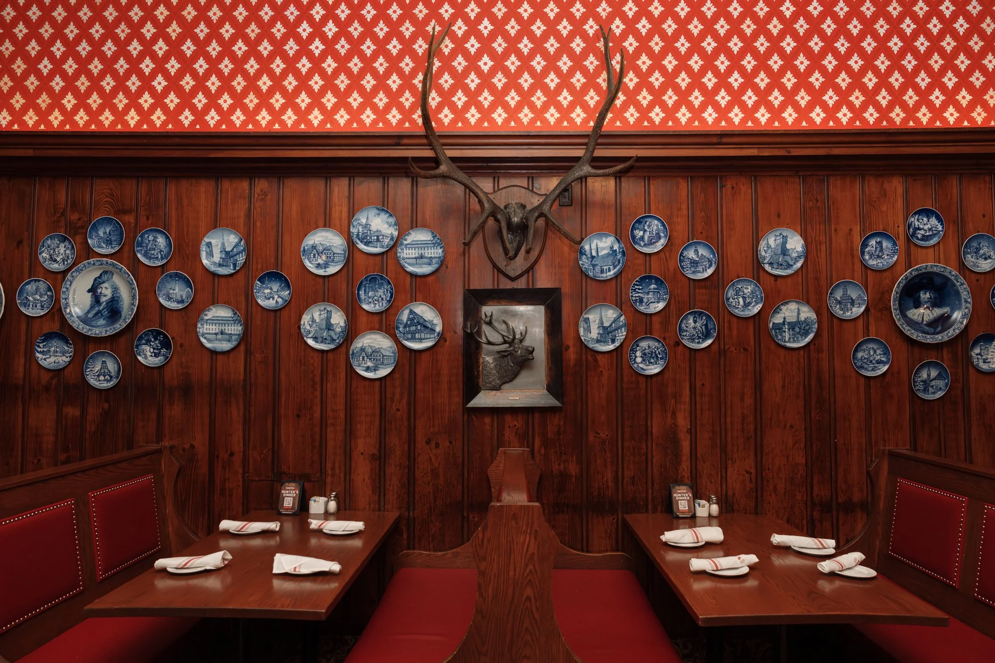 Cozy Dining room at the Student Prince with wooden walls decorated with blue and white plates and mounted animal heads, including a large antlered deer head at the center and wooden booths with place settings.