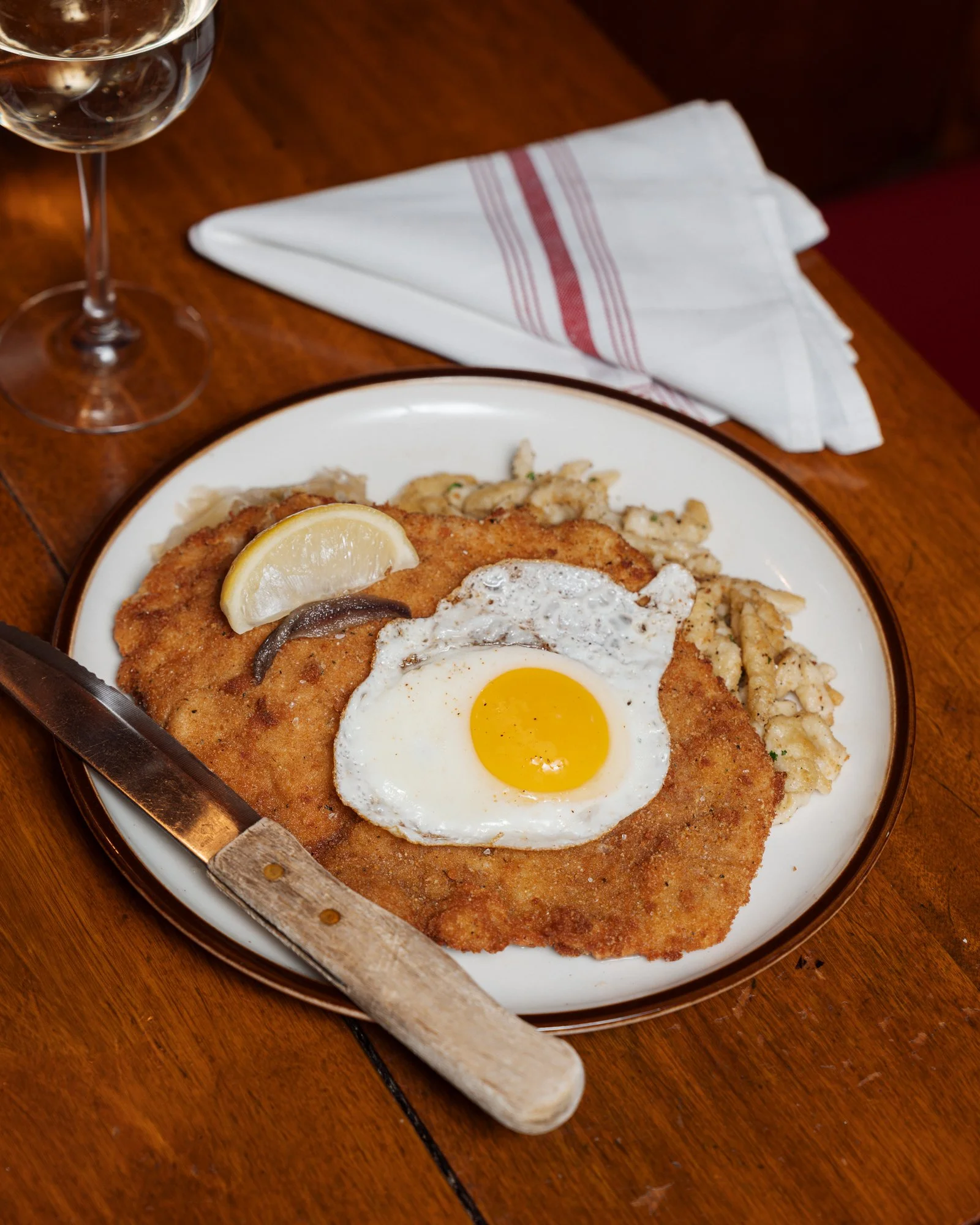 Golden Fried Chicken Schnitzel with a sunny-side-up egg, lemon wedge and anchovy served over Spätzle (a German egg noodle) with a glass of white wine, a knife, and a folded napkin nearby at The Student Prince in Springfield MA
